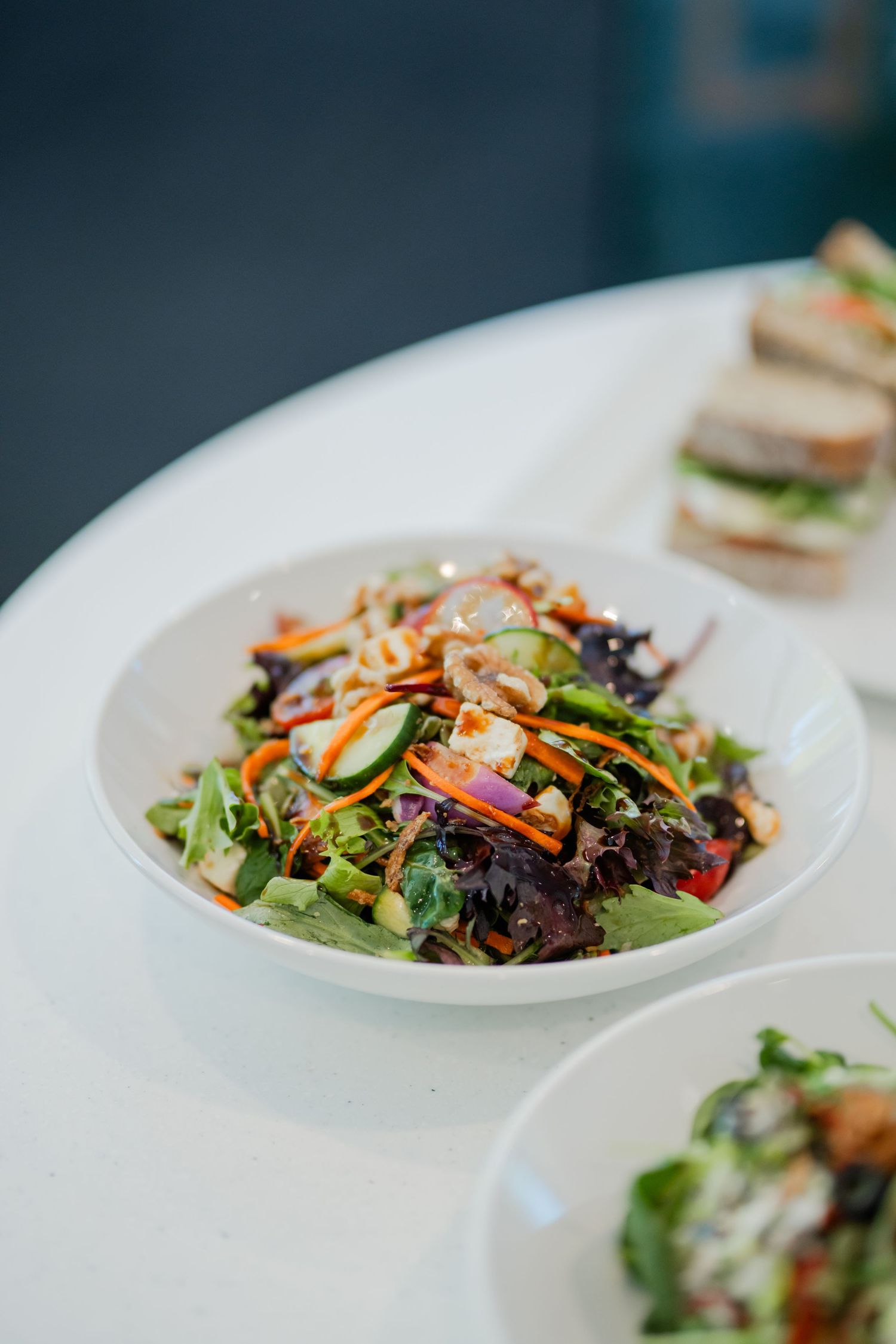 A colorful mixed green salad with carrots, cucumbers, and peanuts in a white bowl on a table.