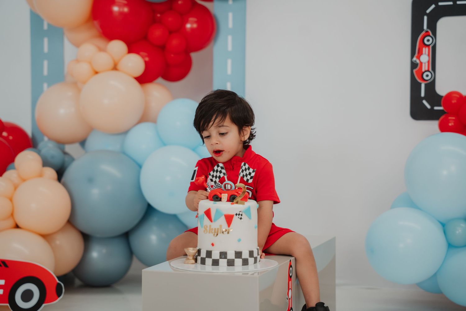 A toddler in red examines his checkered race car birthday cake at a TWO fast balloon-decorated party.