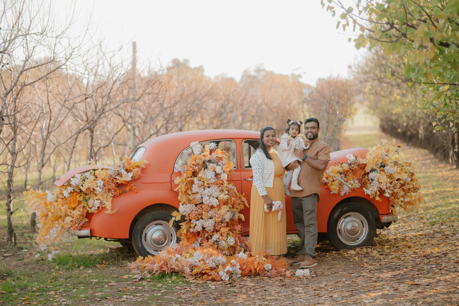 Family of three poses beside a vintage orange car decorated with fall flowers in an autumn orchard.