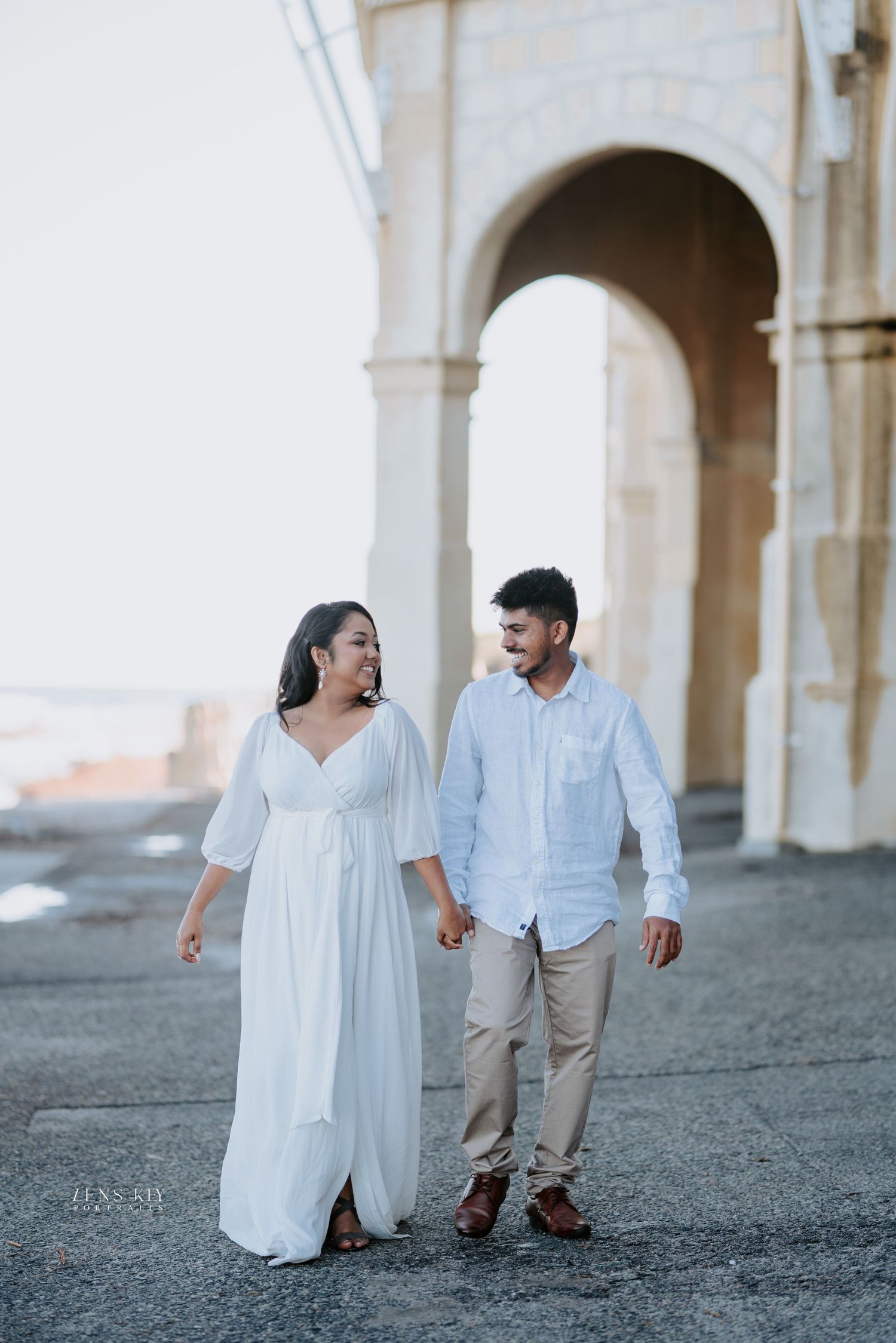 Couple holding hands and walking past a grand stone archway during a romantic engagement photo shoot.