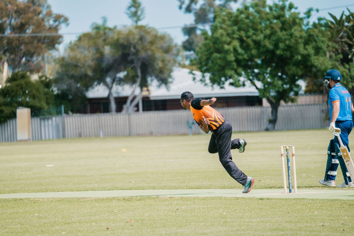 Cricket bowler in orange kit delivers ball near wickets during match.