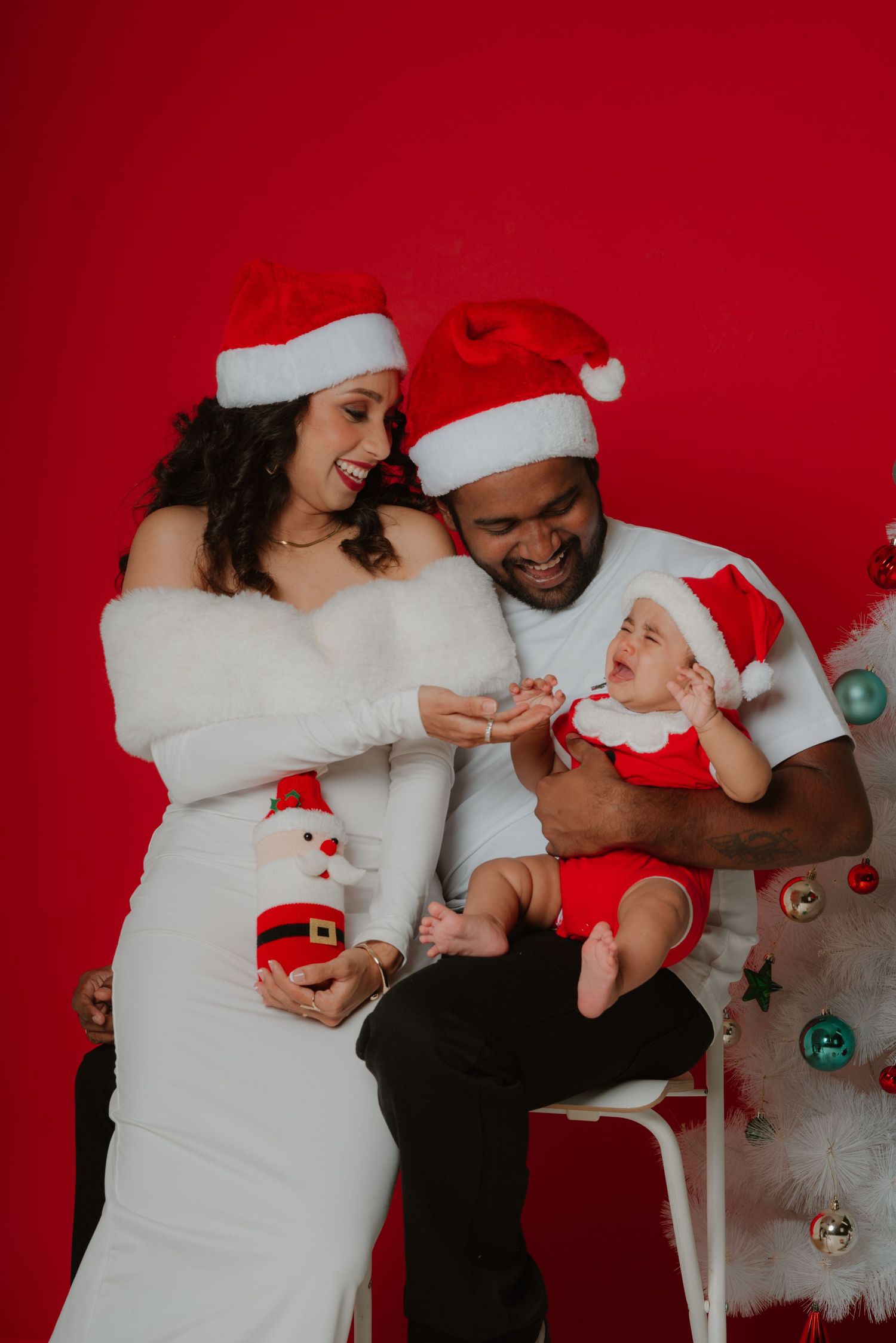 Laughing family in Santa hats holding a crying baby in holiday outfit beside a decorated white Christmas tree.
