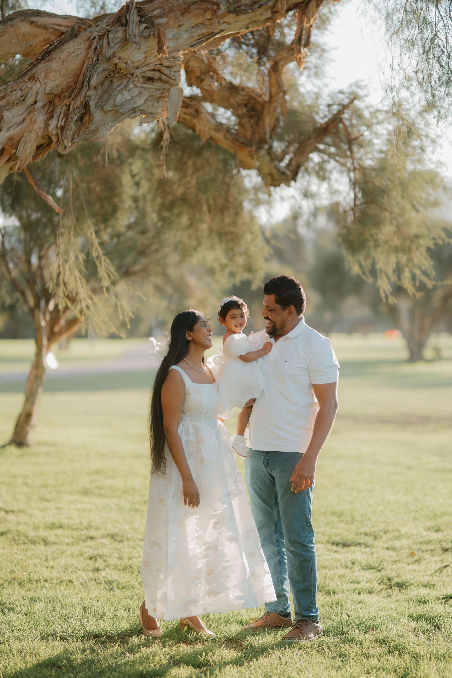 Happy family of three standing under a tree in a sunny green park, smiling together.