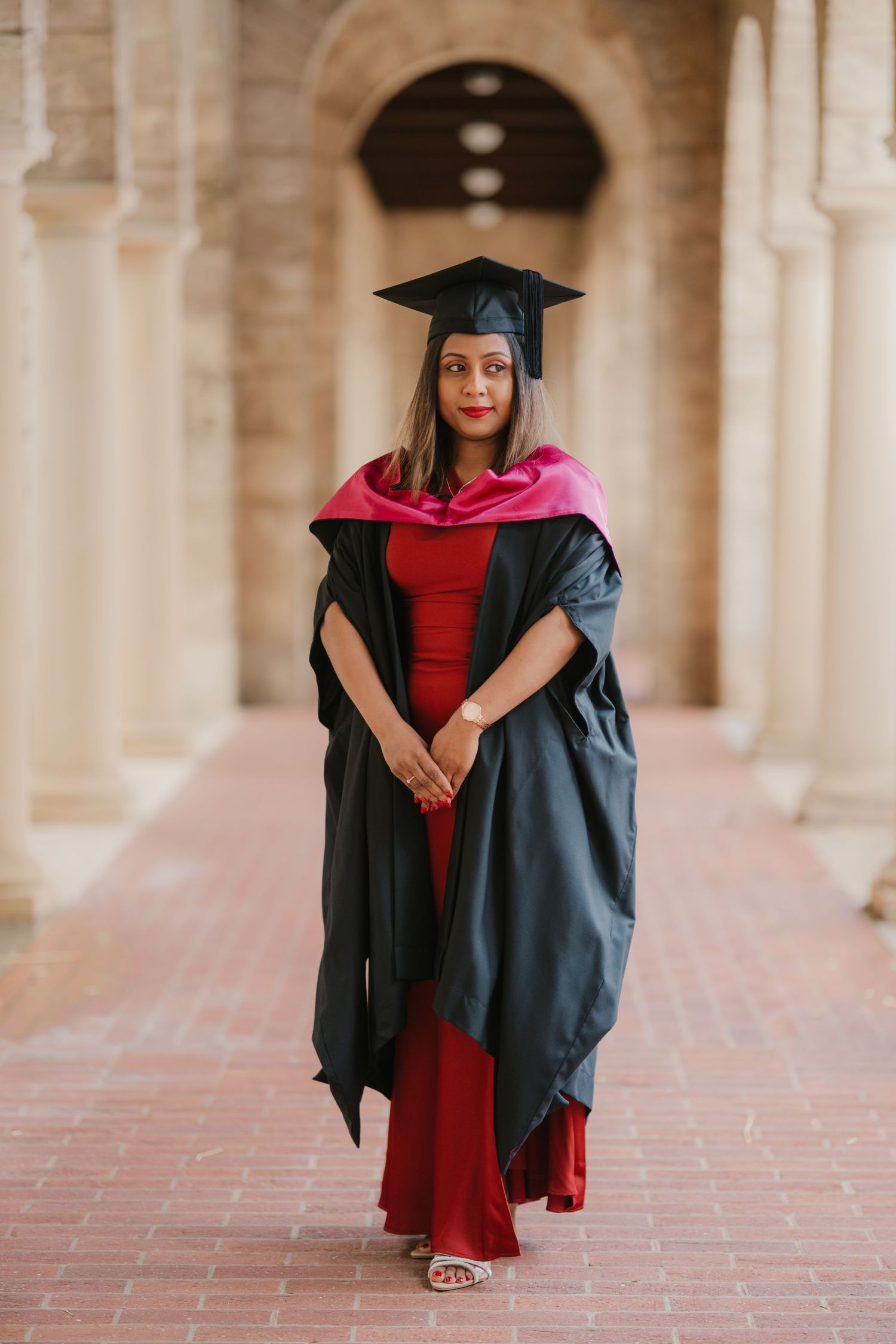 Female graduate in red dress and black gown poses in university archway corridor.