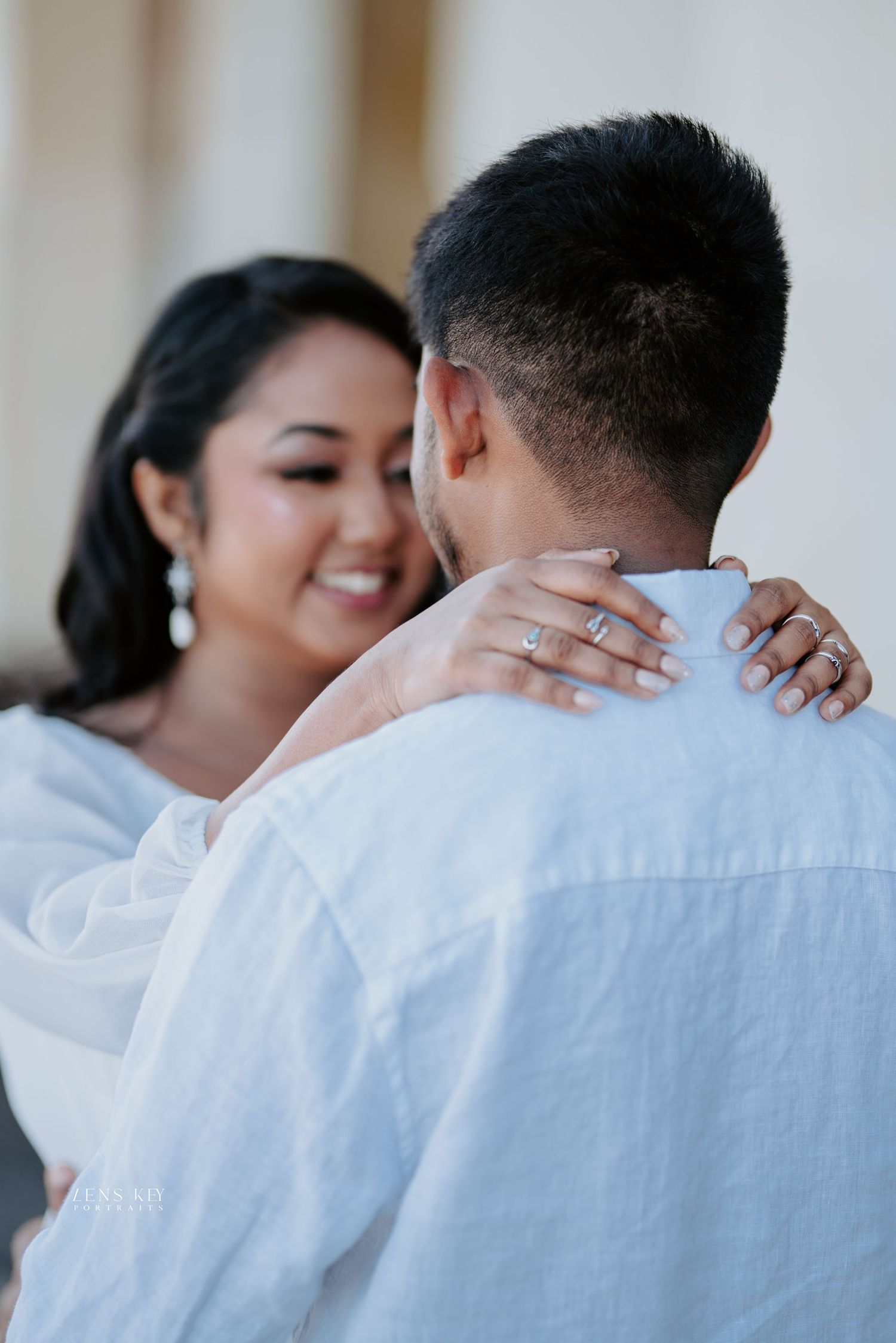 Smiling bride in white dress embracing groom from behind, showcasing elegant rings and drop earrings.