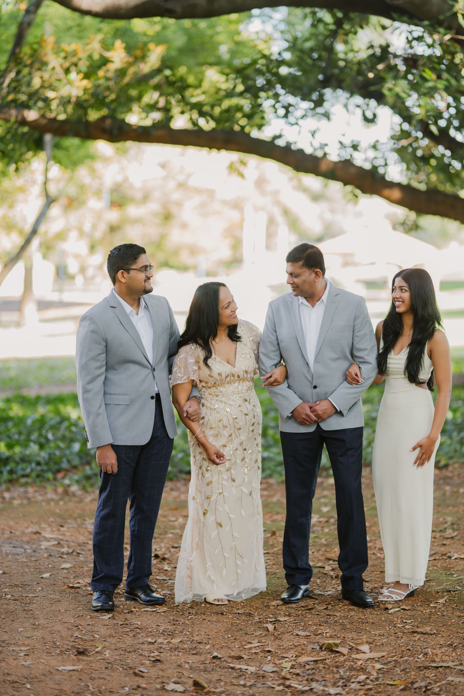 Family of four laughs together on a sunlit dirt path surrounded by green trees in casual formal wear.