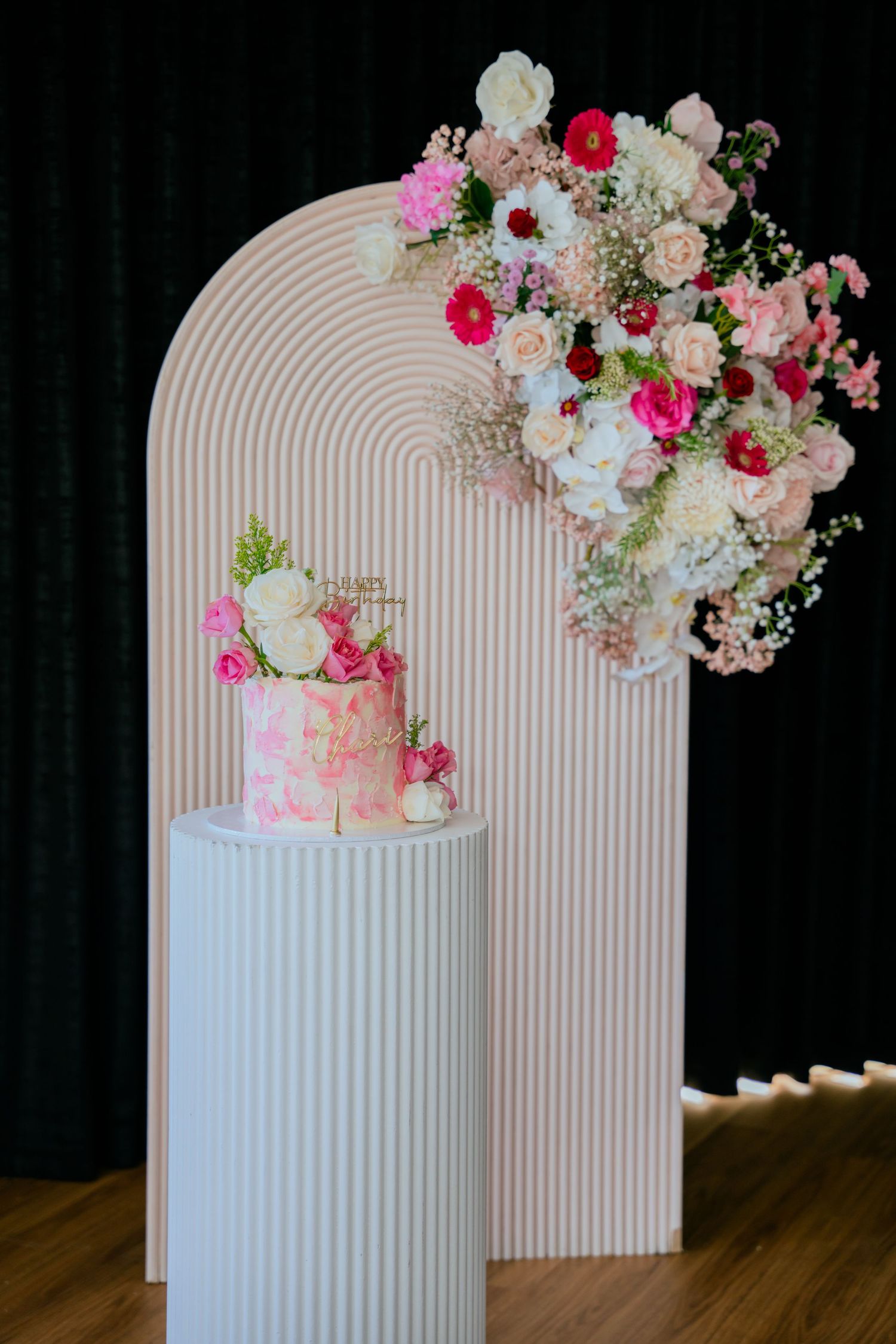 Elegant pink floral birthday cake on ribbed pedestal with arch backdrop and flower arrangement.