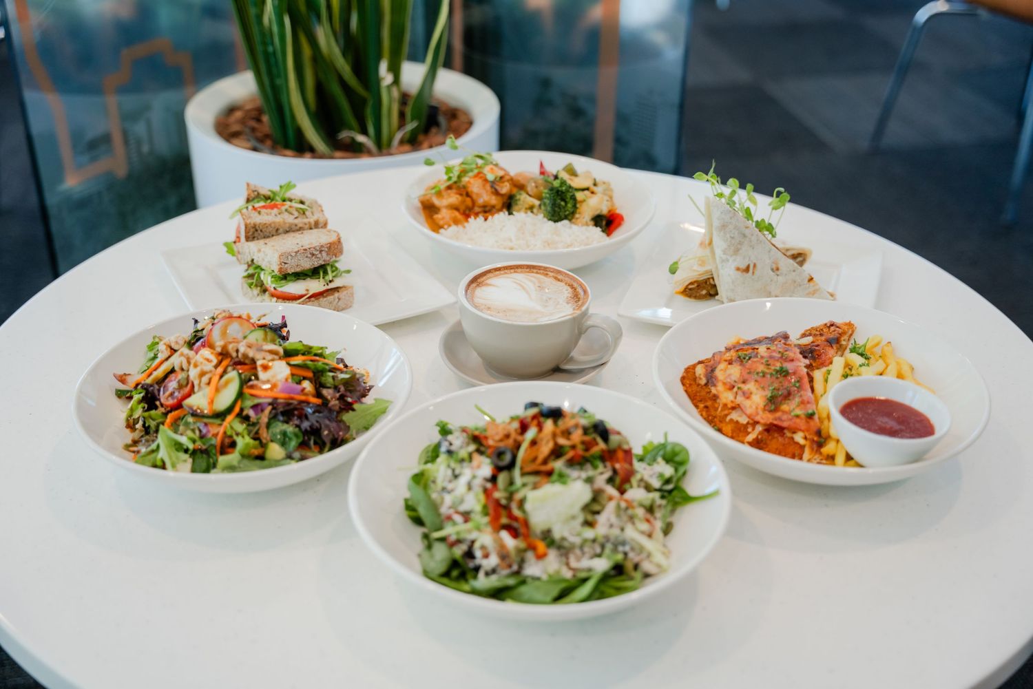 Overhead view of a spread of salads, pasta, sandwiches, and lattes on a white round table.