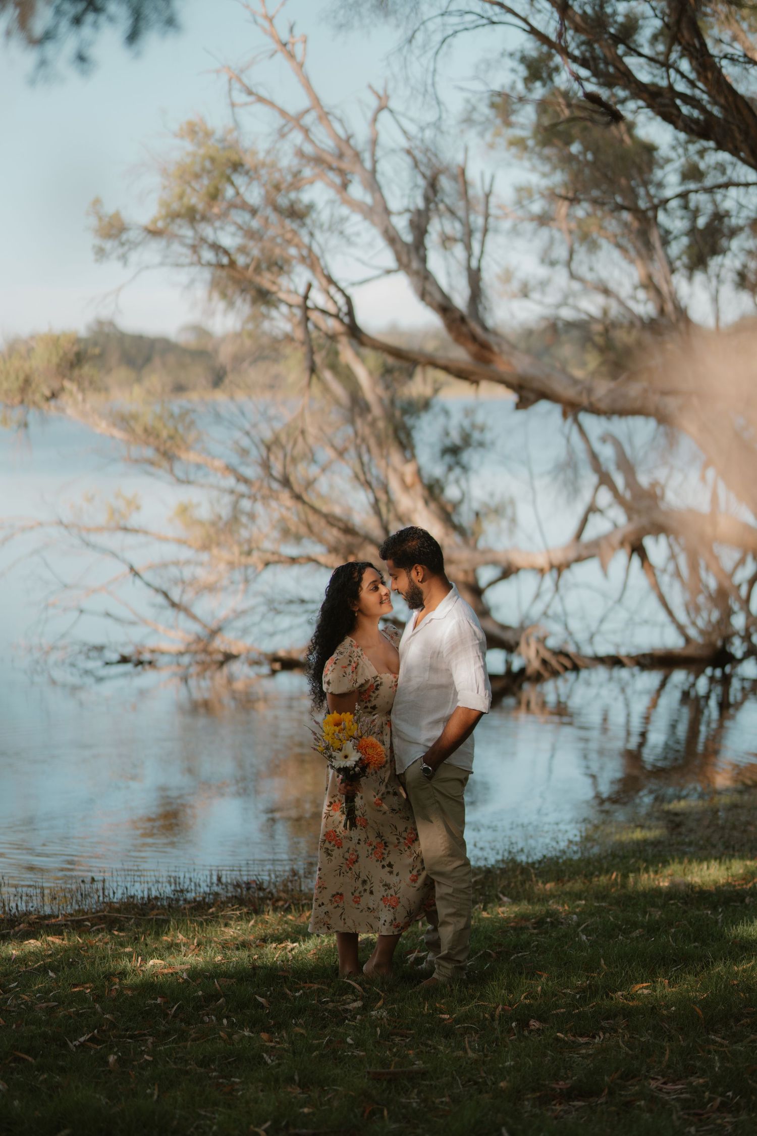 A couple shares an intimate moment near a serene lake with golden autumn trees in the background.