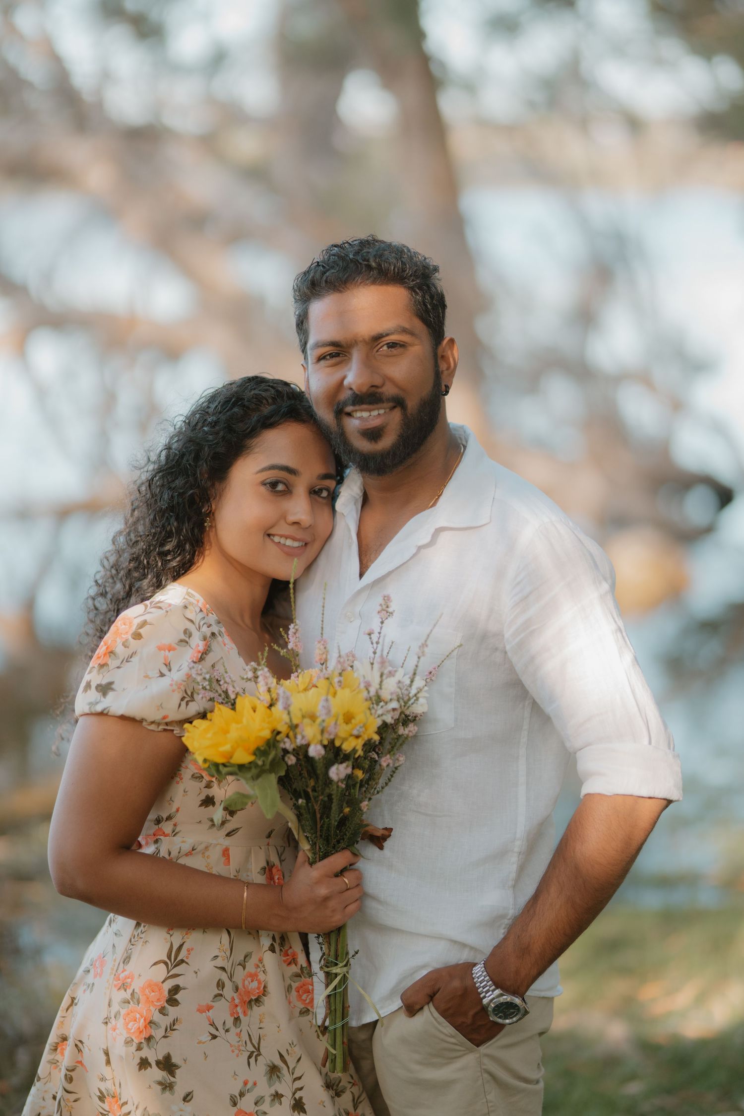 Smiling couple posing outdoors holding yellow wildflowers with soft bokeh tree background.