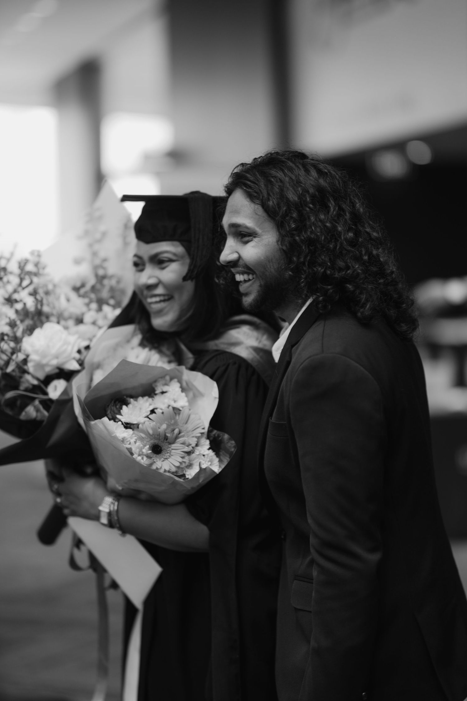 Black and white photo of a graduate in cap and gown laughing with a man holding flowers.