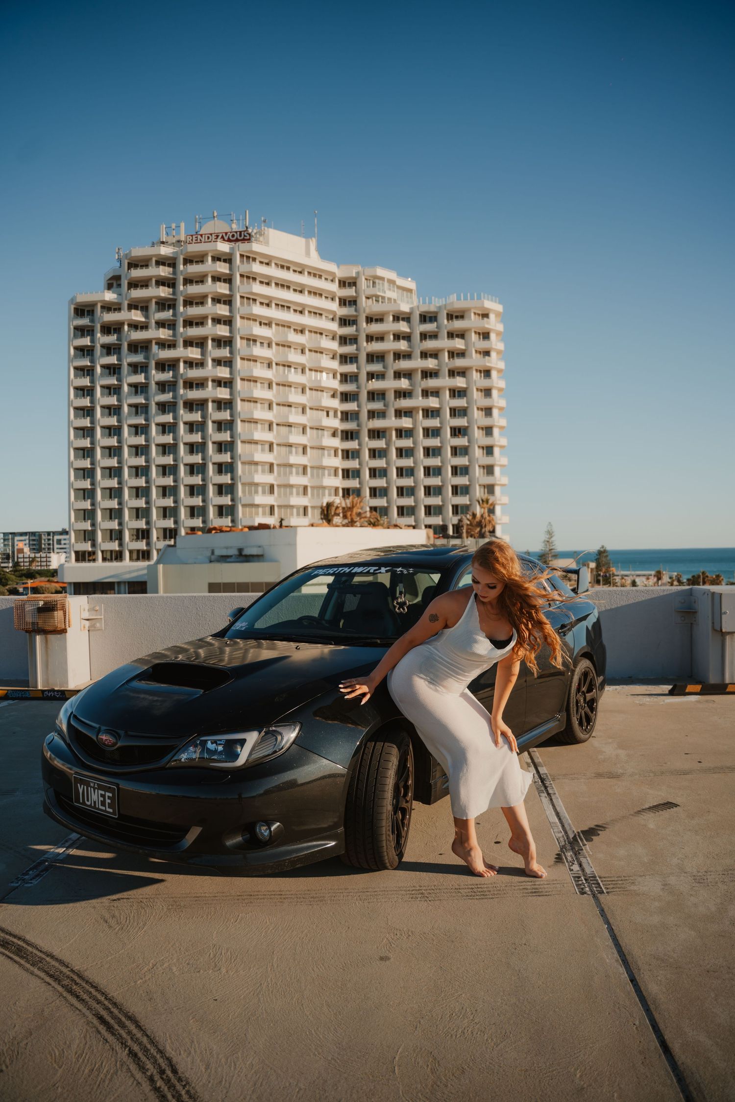 Woman in white dress leaning on black Subaru WRX on rooftop with high-rise building backdrop.