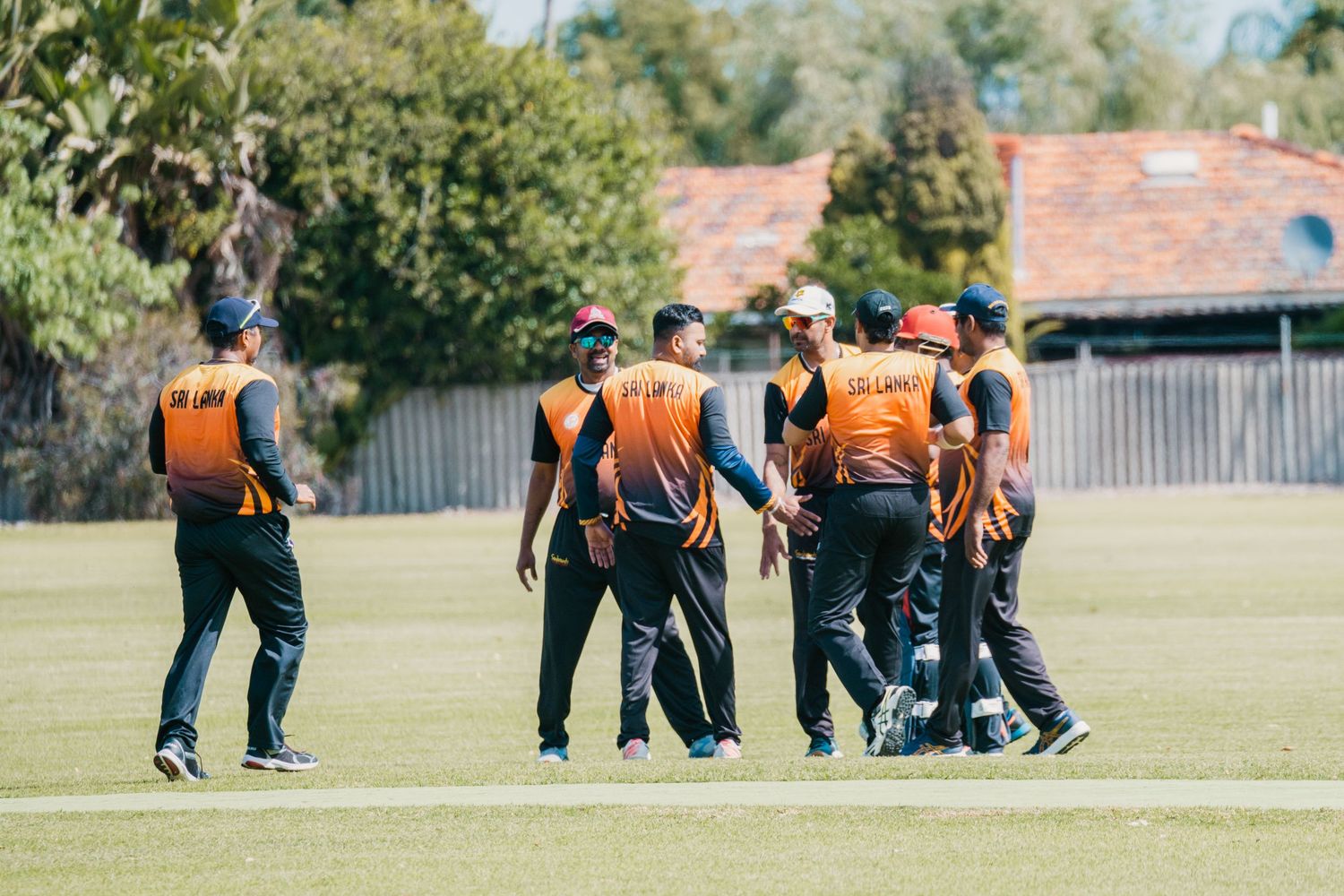 Cricket team in orange uniforms celebrating together on a green field during a sunny match.