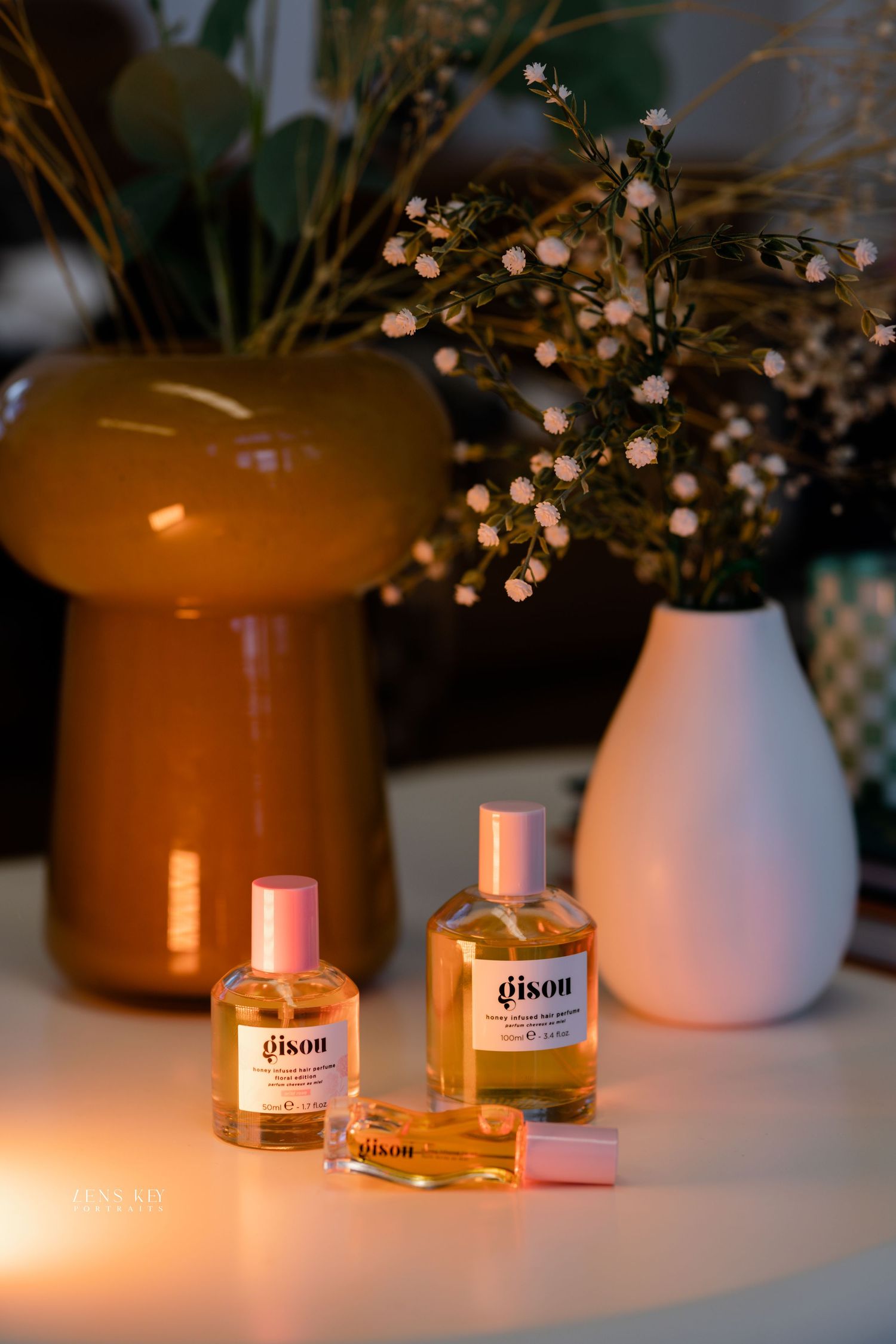 Three Gisou honey infused hair perfume bottles with pink caps displayed near amber vase and white floral arrangement.