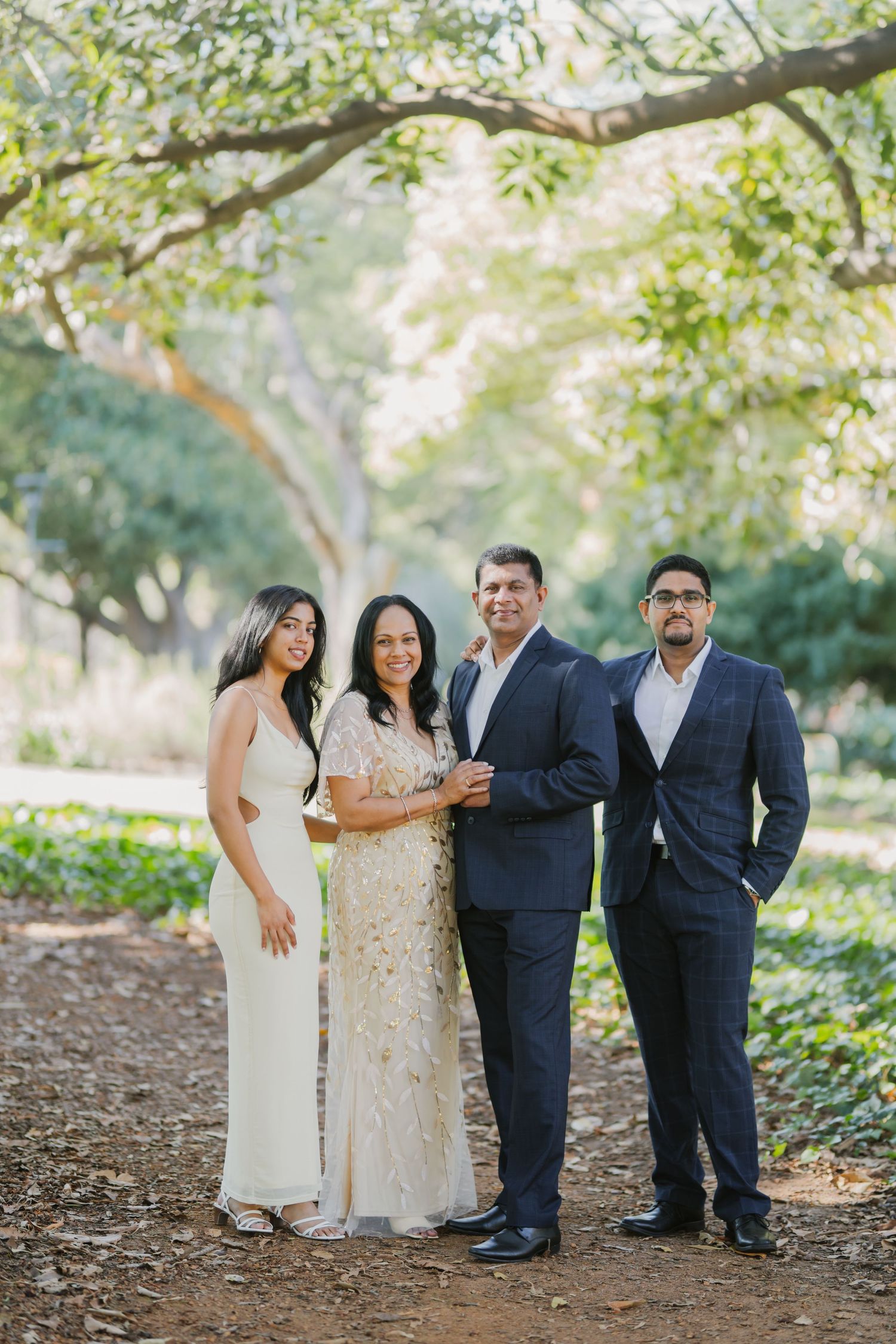 Elegant family of four smiles together under lush trees, dressed in formal attire for a portrait session.