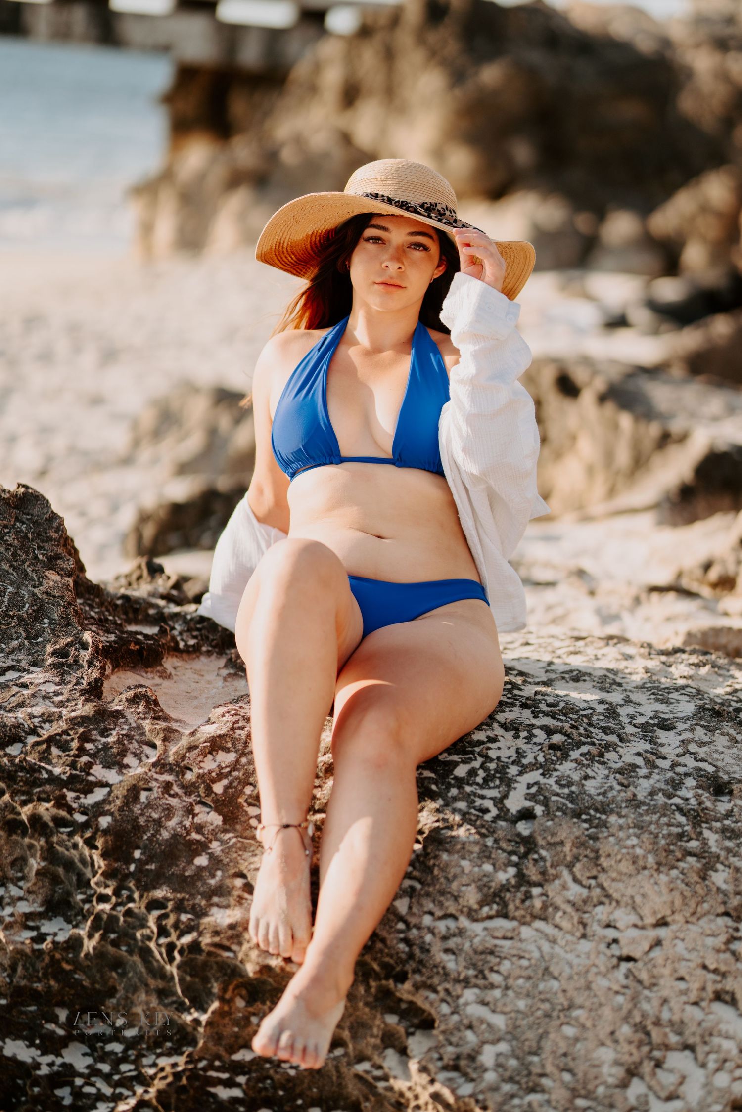 A woman in a blue bikini and white shirt sits on coastal rocks wearing a wide-brim straw hat.