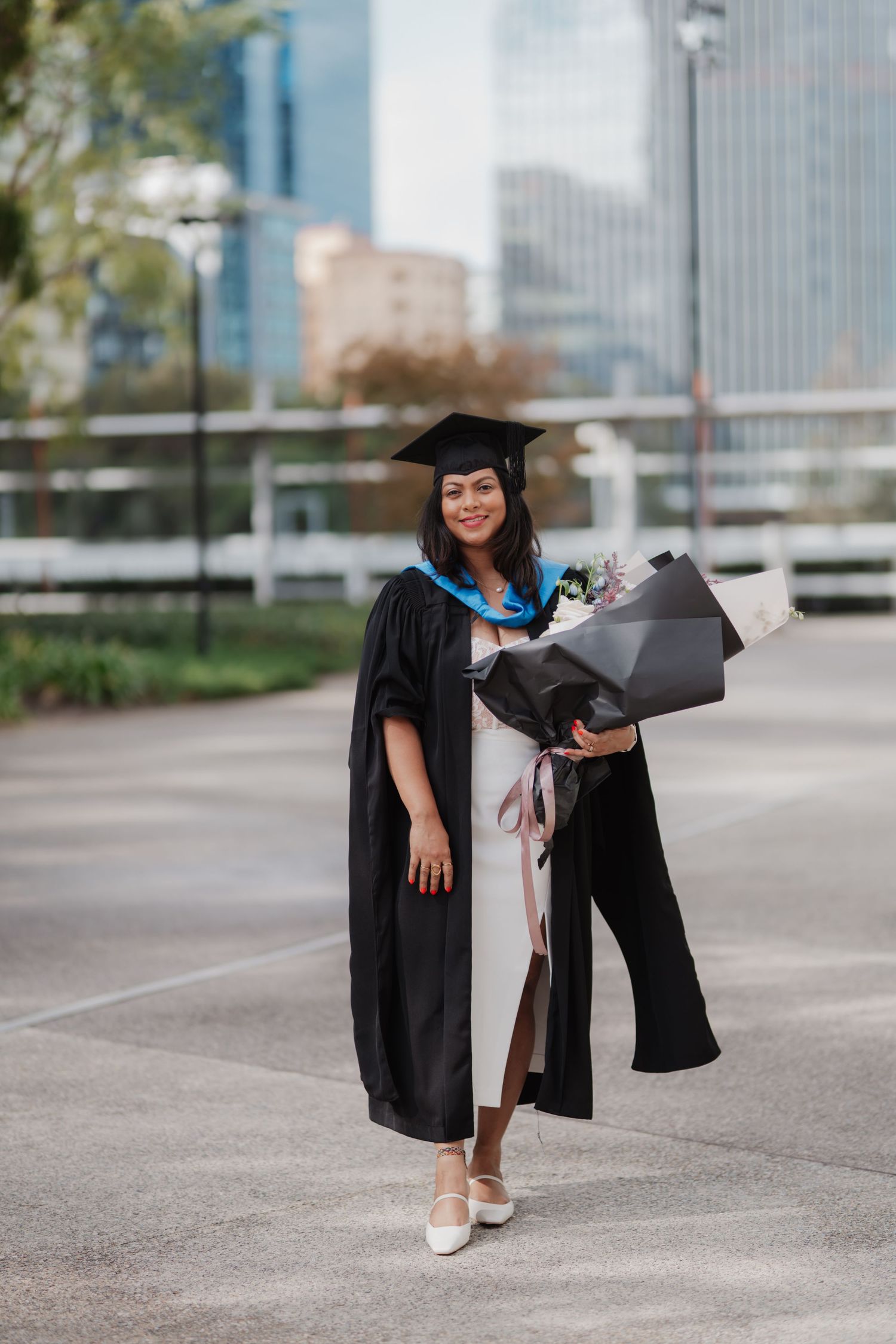 Smiling graduate in black cap and gown holds flower bouquet with city skyline background.