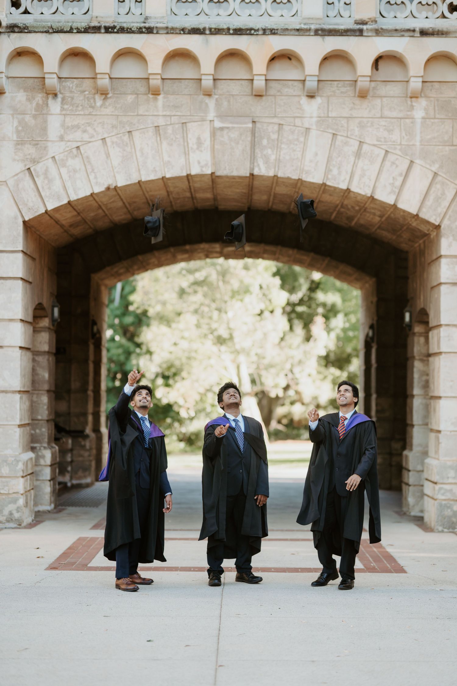 Three graduates in black robes tossing caps under stone archway at university campus.