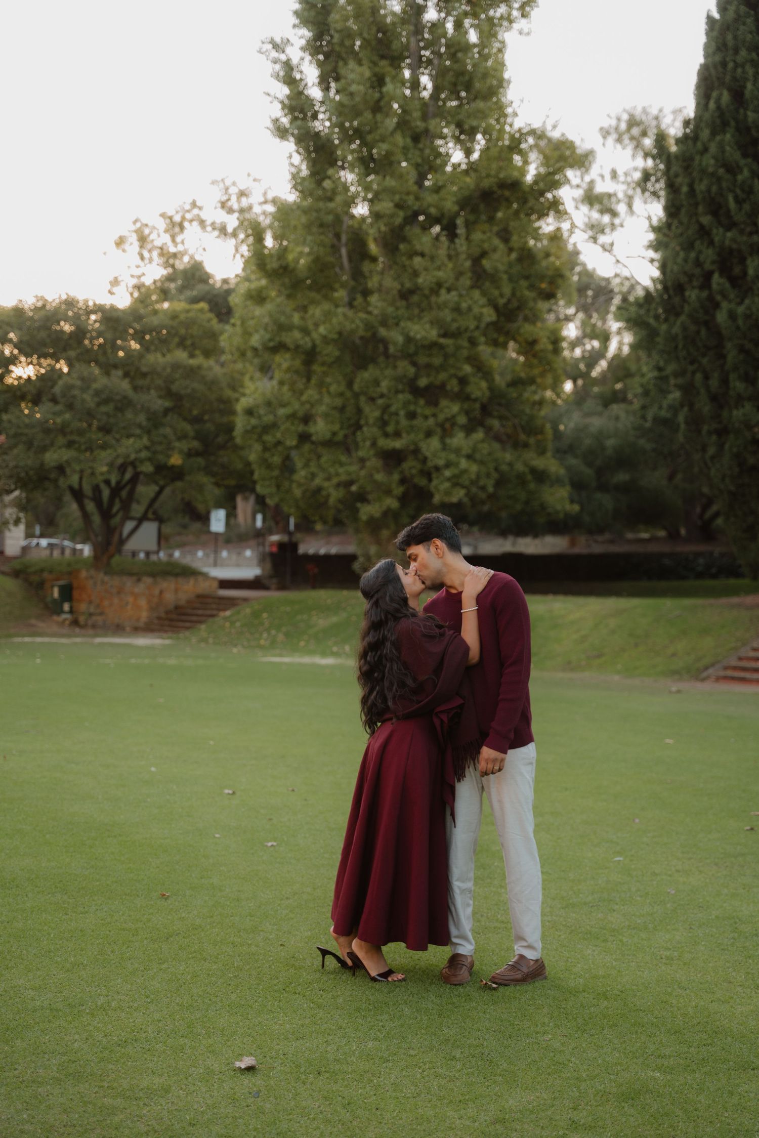 Romantic couple shares a kiss on lush green lawn surrounded by tall trees at dusk.
