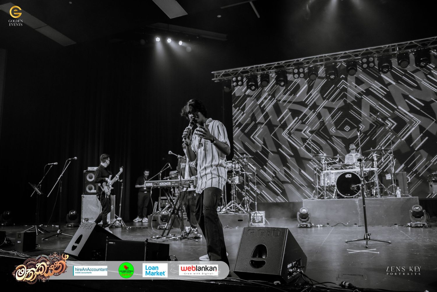 Black and white wide shot of band performing on stage with dramatic geometric backdrop.