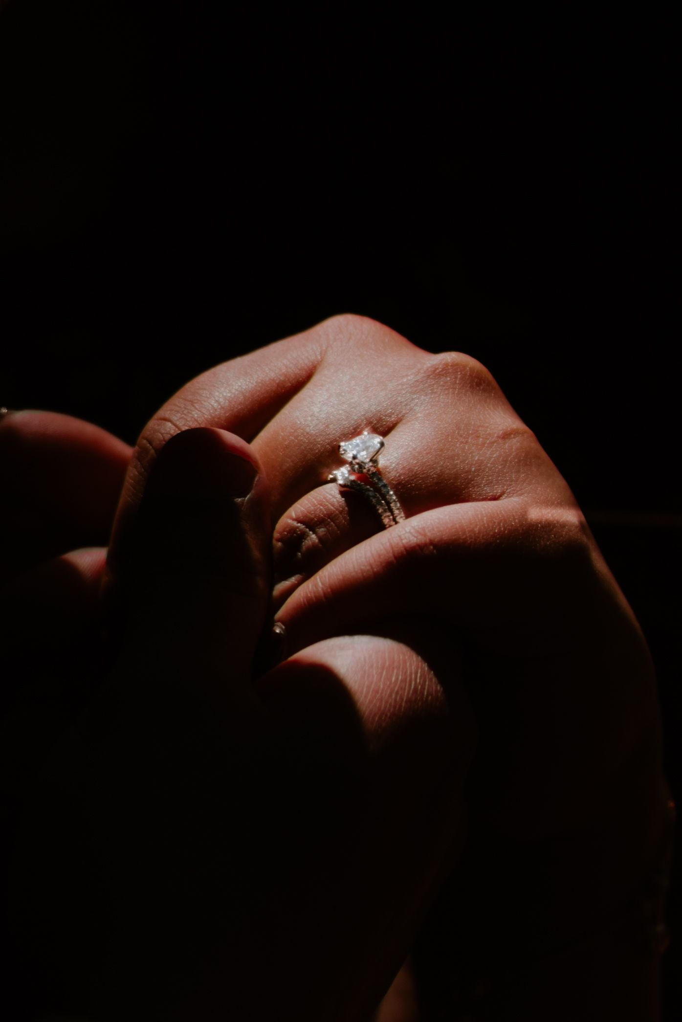 Close-up of diamond engagement ring on hand dramatically lit against dark background.