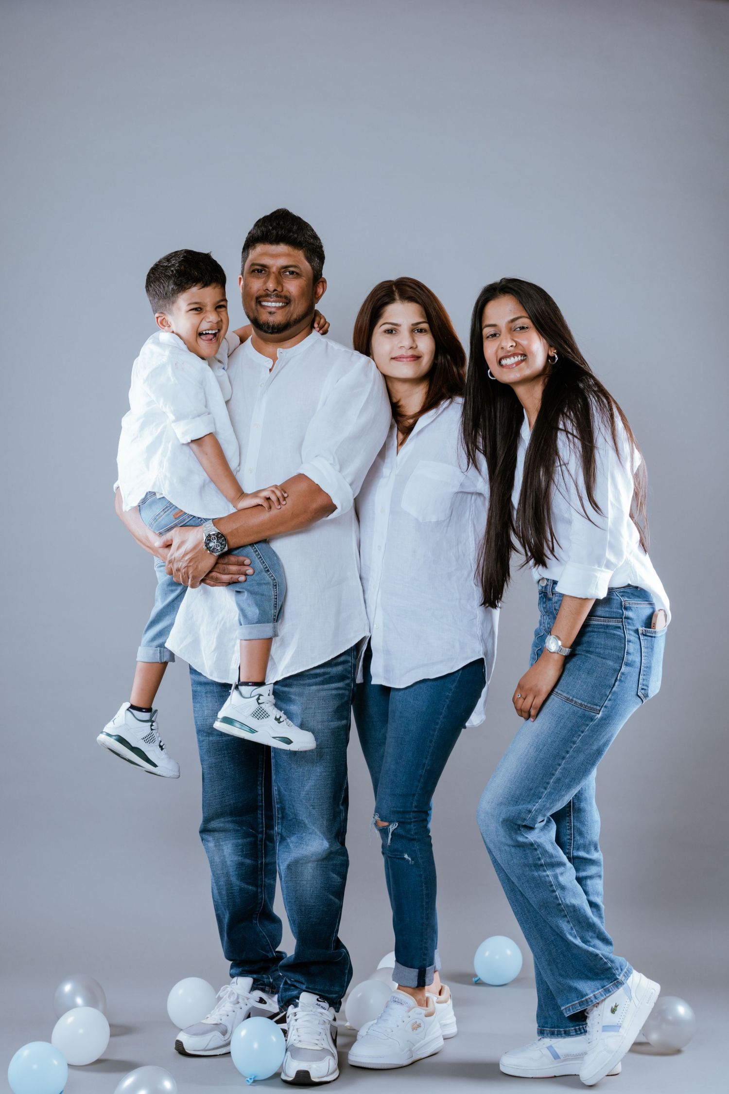 Happy family of four in white shirts and jeans pose with balloons on gray background.