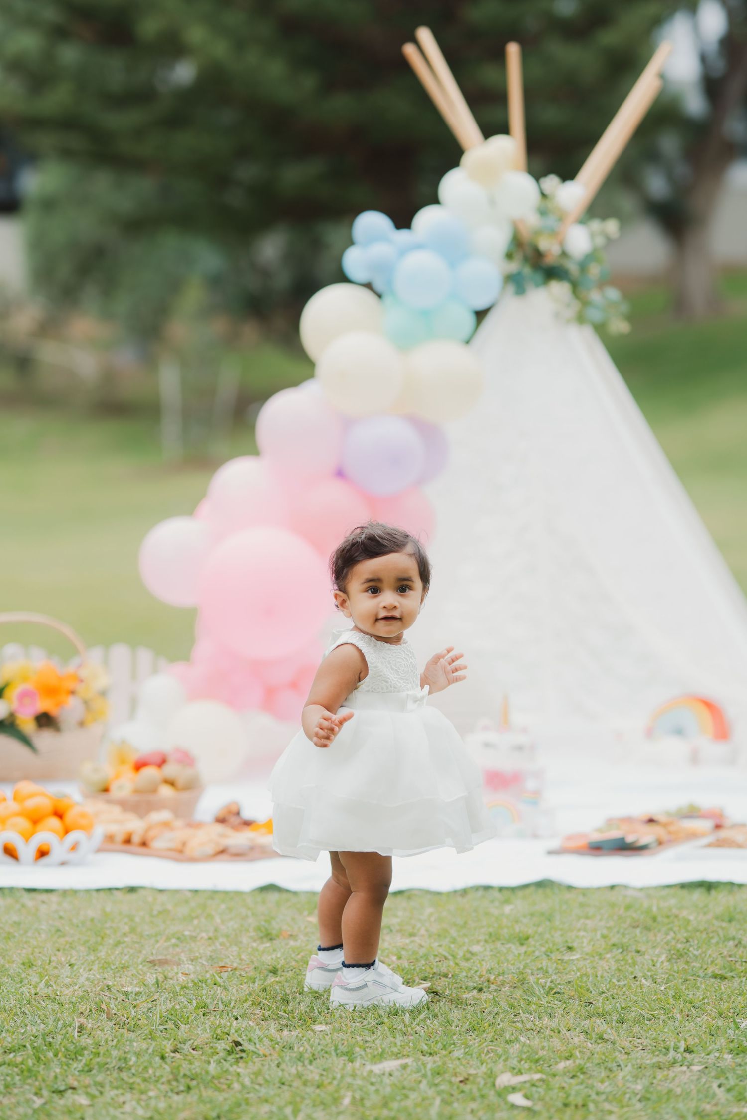 Baby girl in white dress standing on grass at a colorful unicorn first birthday party.