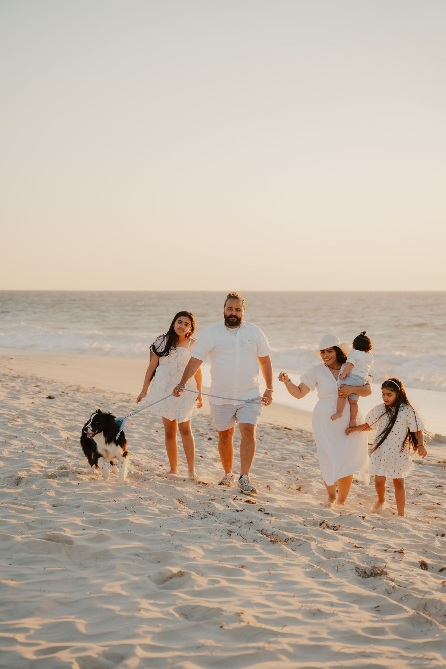Happy family in white outfits walking on beach at sunset with a border collie dog.
