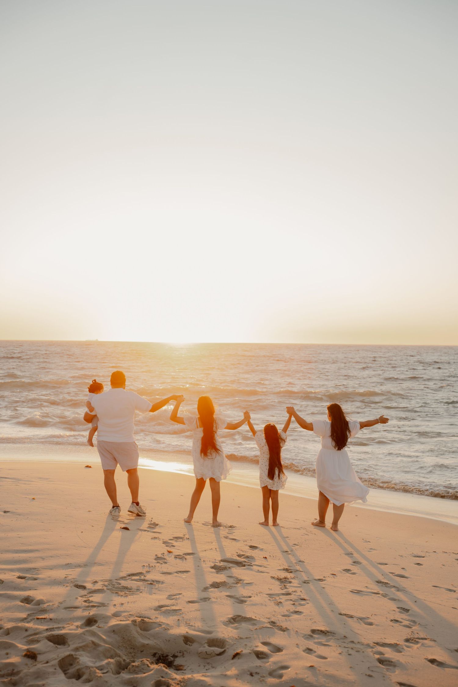 Family of five in white clothing raising arms joyfully on beach during golden sunset.