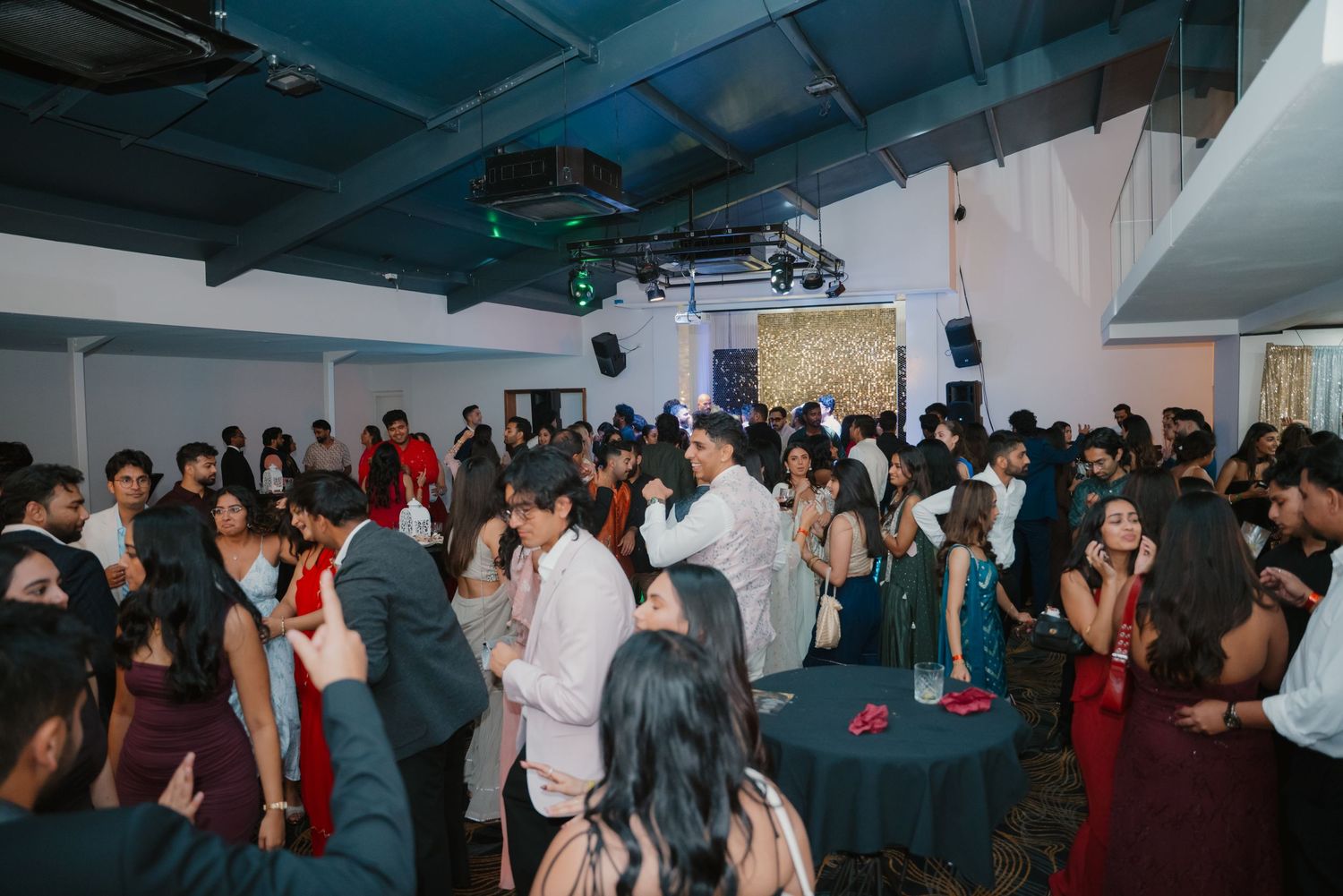 Crowded indoor Indian wedding reception with guests mingling and dancing under blue ambient lighting.