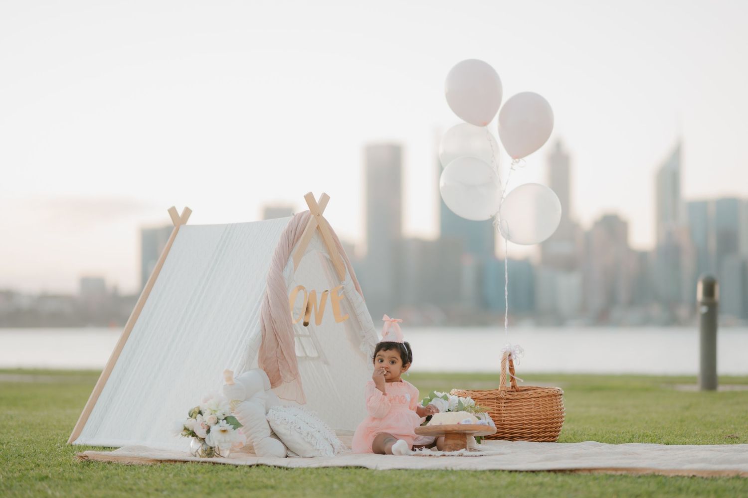 Baby girl in pink dress sitting by a white tent with balloons and city skyline backdrop.