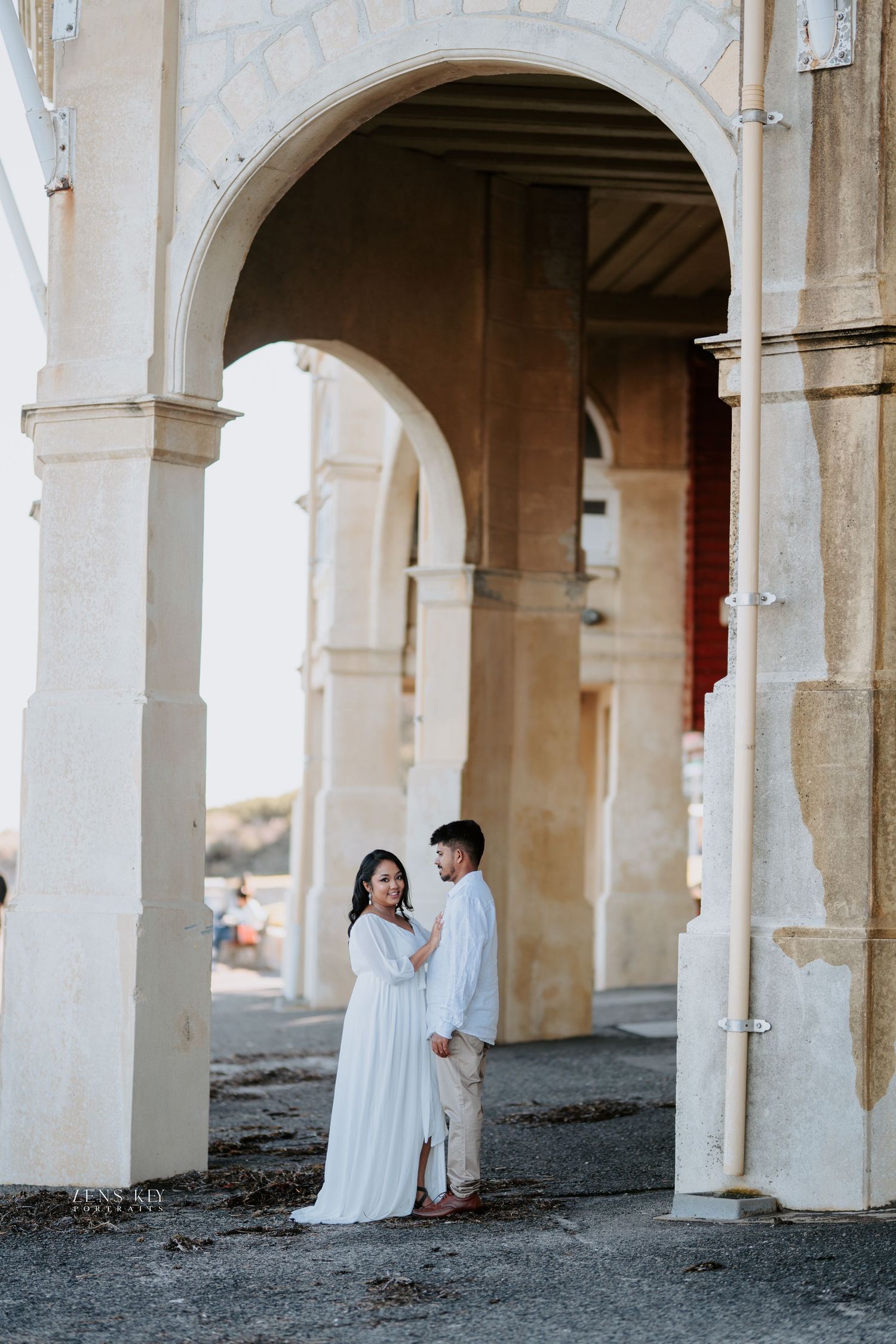 Couple sharing a tender moment beneath elegant white arched columns during an outdoor engagement session.