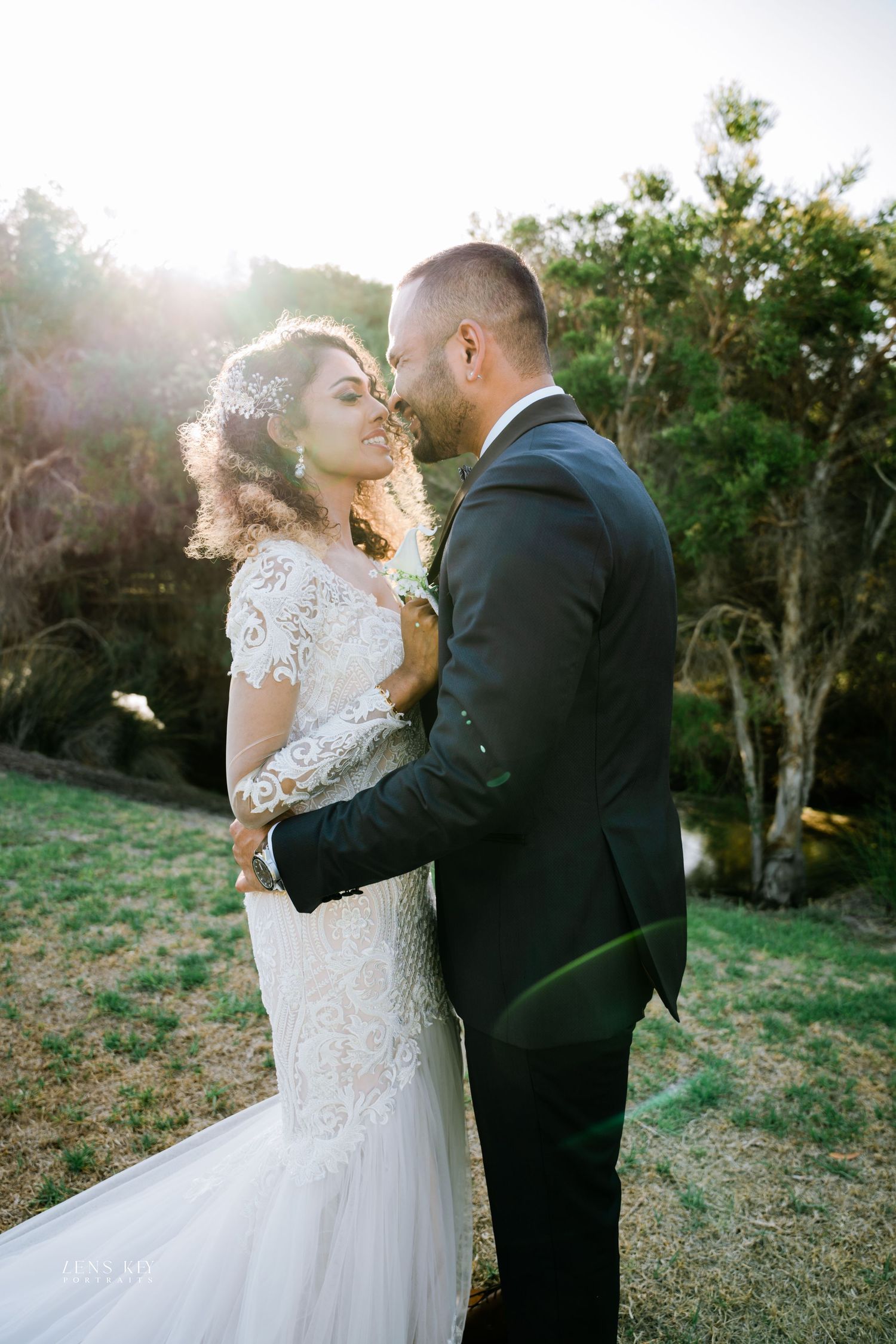 Bride and groom share romantic moment at golden hour sunset, bride in lace mermaid gown embraced by groom in dark suit.