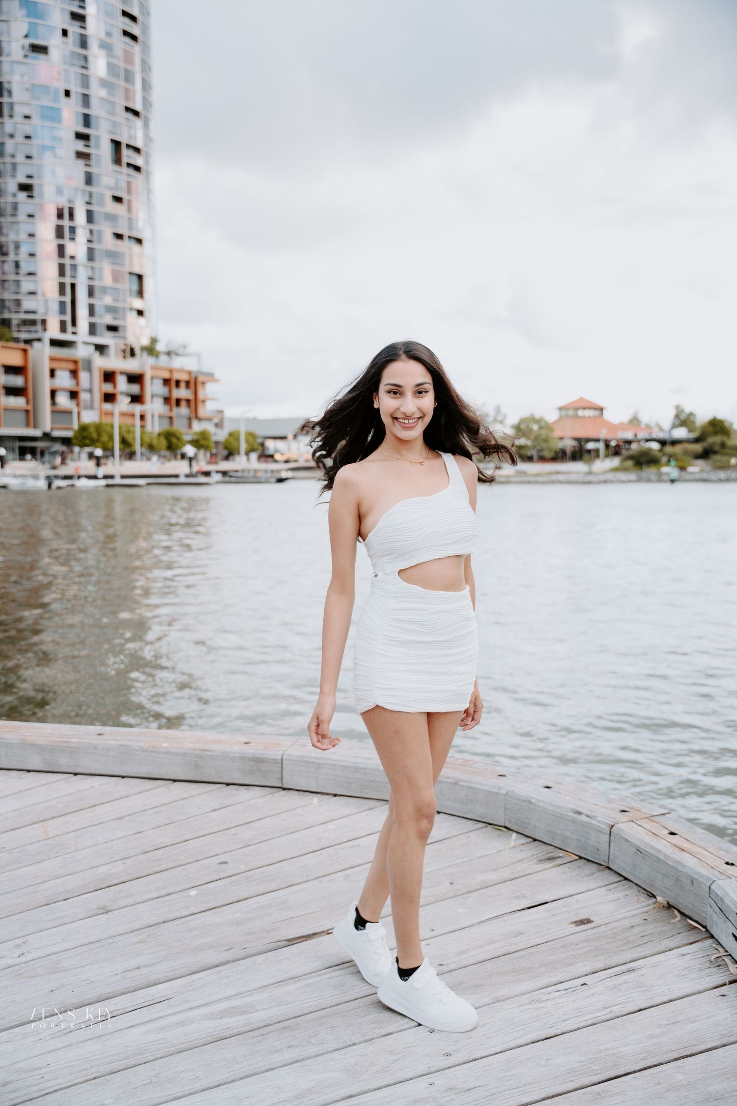 Smiling woman in white cutout one-shoulder mini dress standing on waterfront boardwalk.
