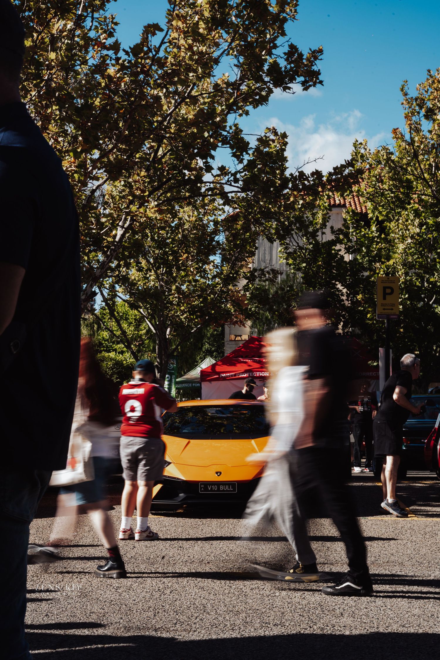 Blurred pedestrians walking past a yellow sports car parked on a sunny tree-lined street.