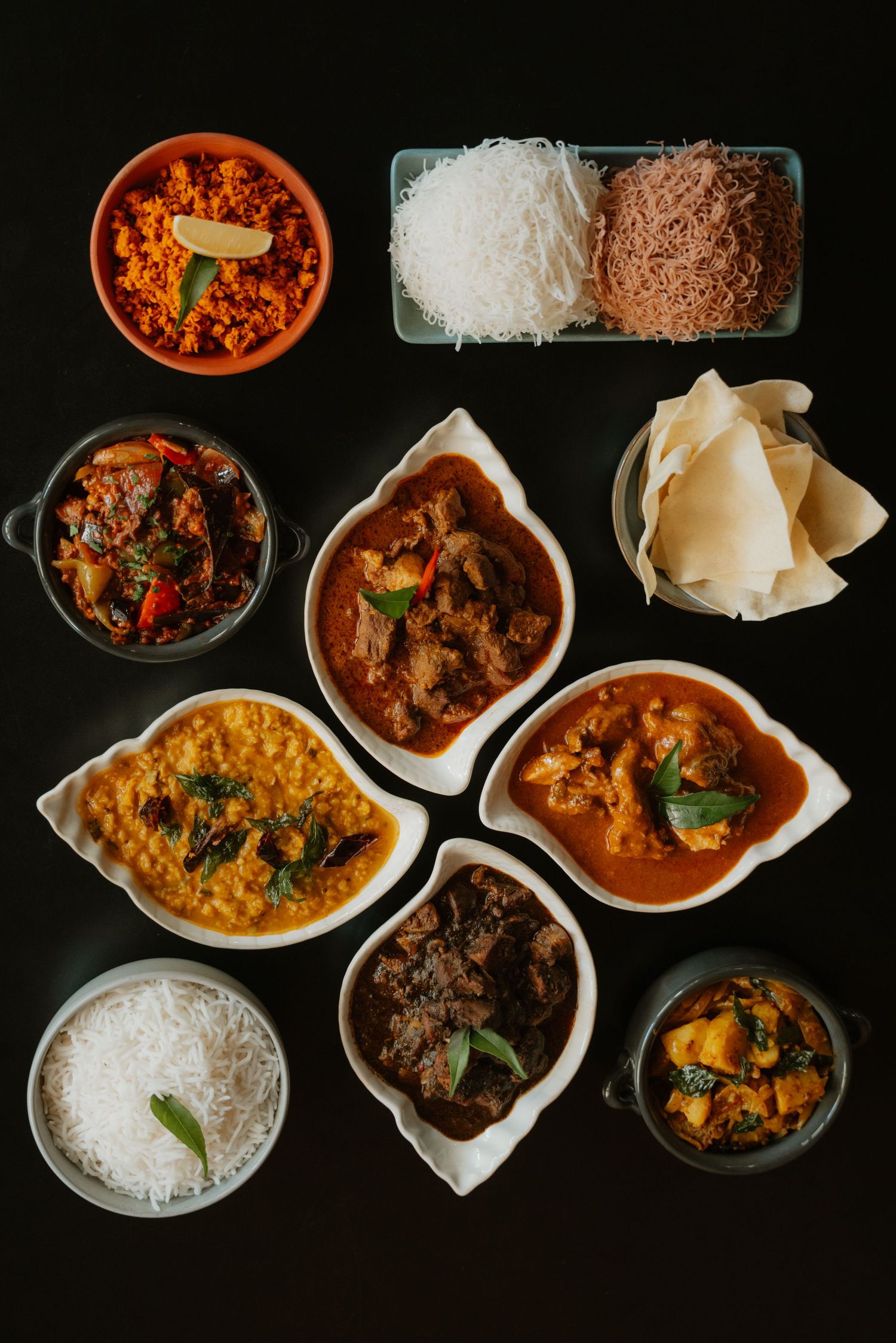 Overhead view of traditional Sri Lankan meal with curries rice string hoppers and papadum on black surface.