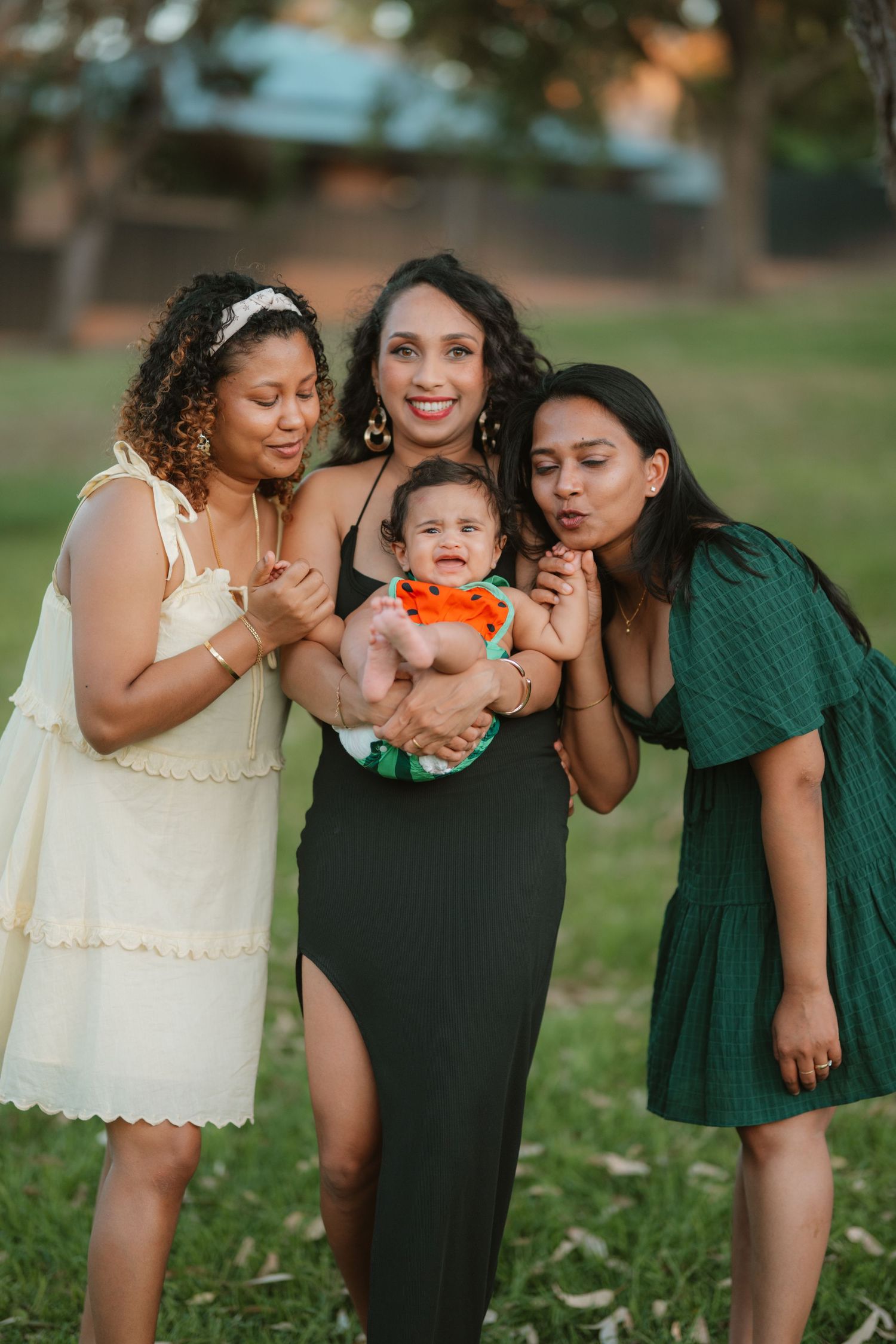 Four women outdoors at golden hour, three adults in dresses surrounding a woman holding a baby in a park.