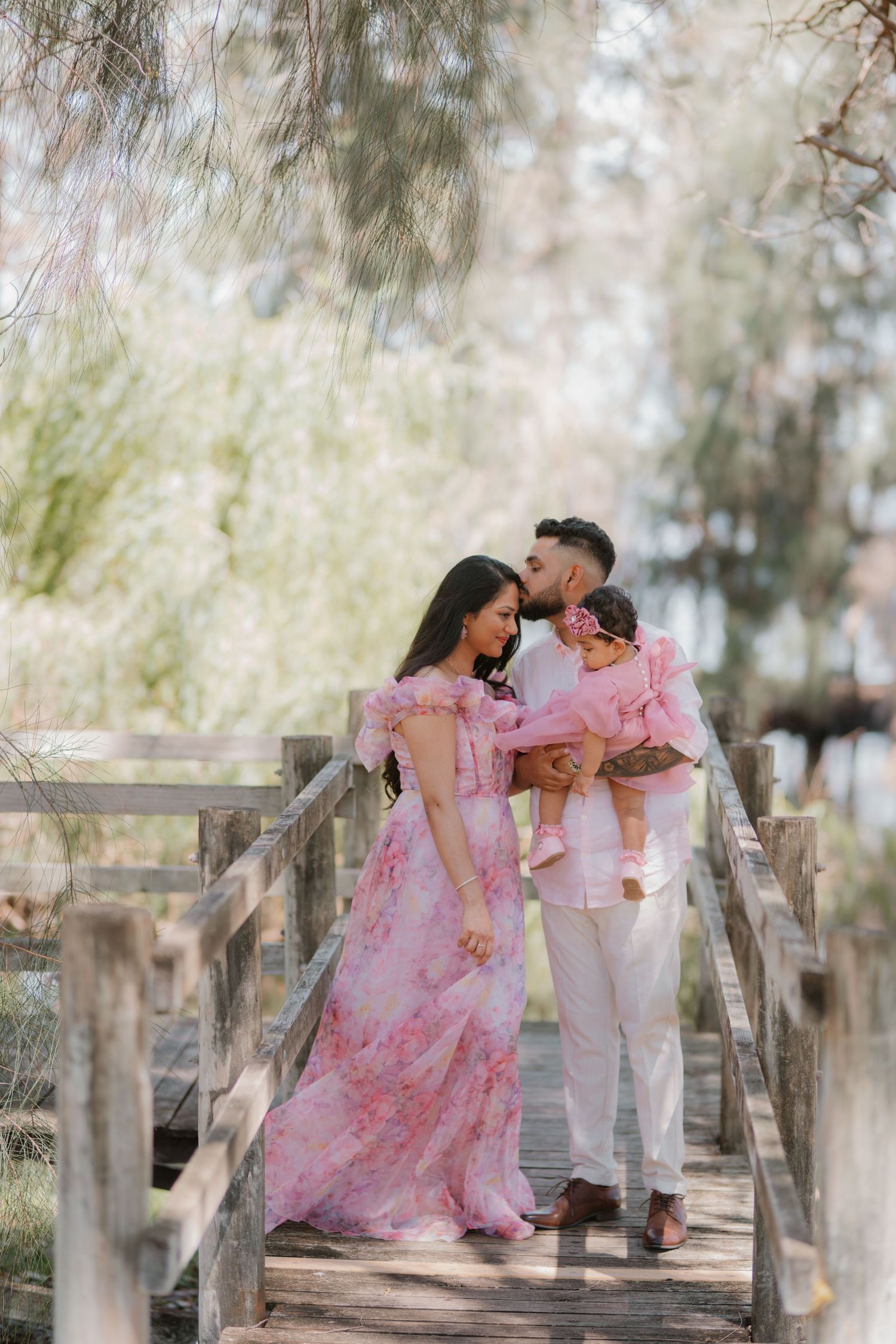 A family in coordinating pink outfits poses on a wooden bridge surrounded by trees.