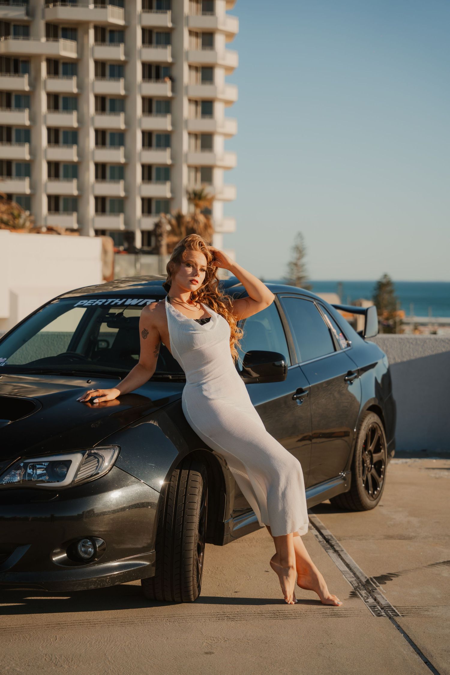 Blonde woman in white midi dress posing barefoot against a black Subaru WRX on a rooftop.