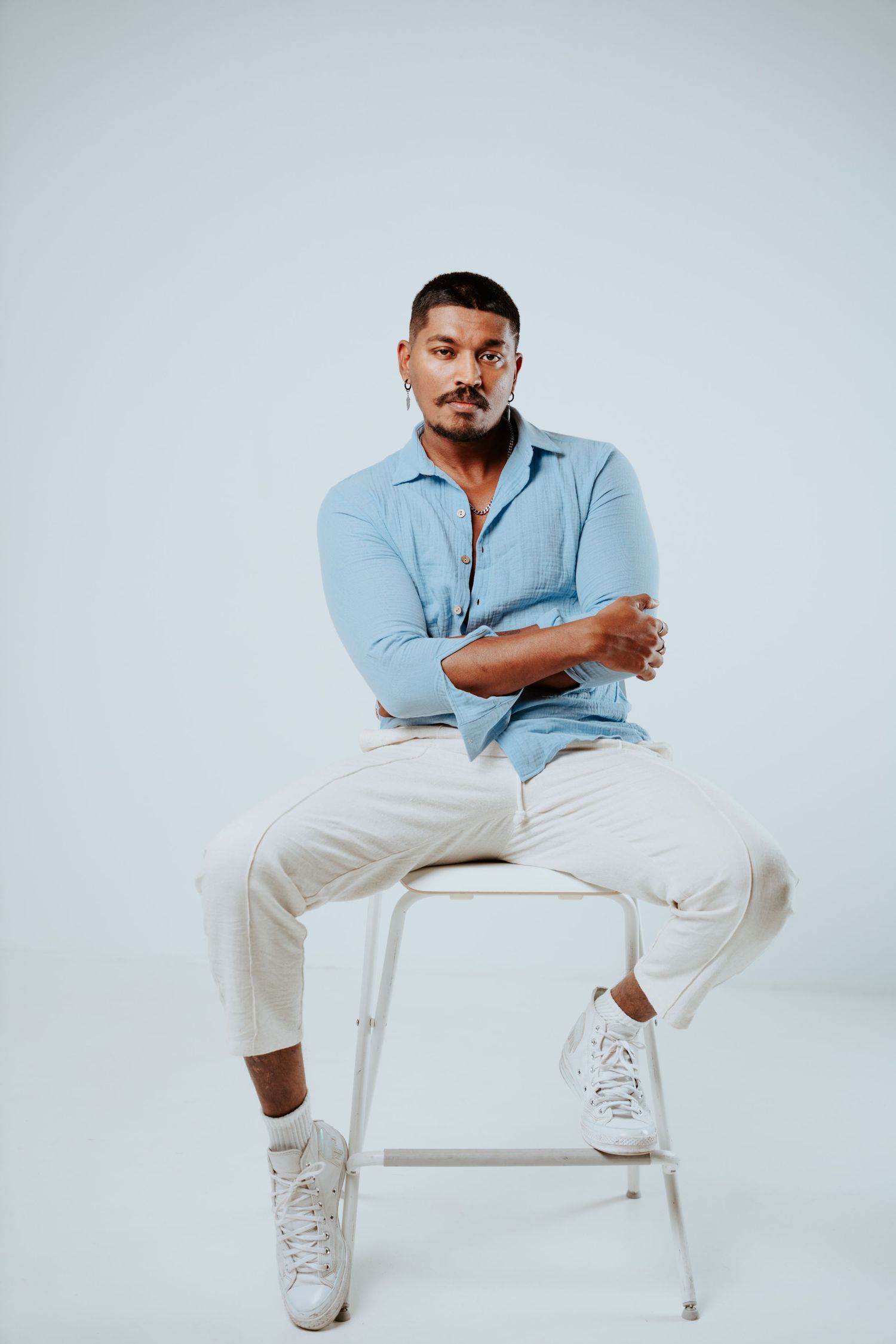 Man in blue shirt and white pants sits cross-legged on white stool in minimalist studio.