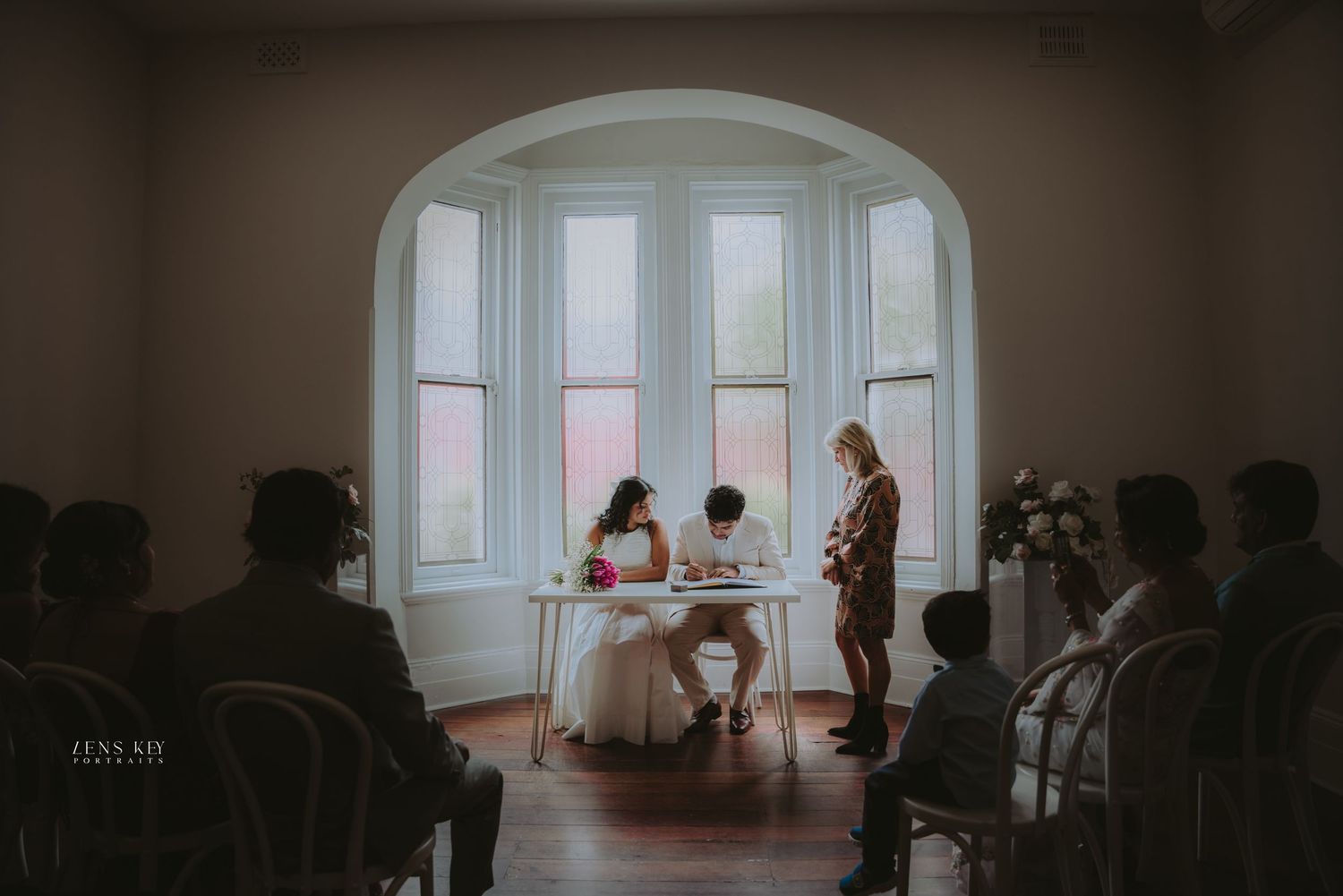 Intimate indoor wedding ceremony at bay window with couple signing marriage documents.
