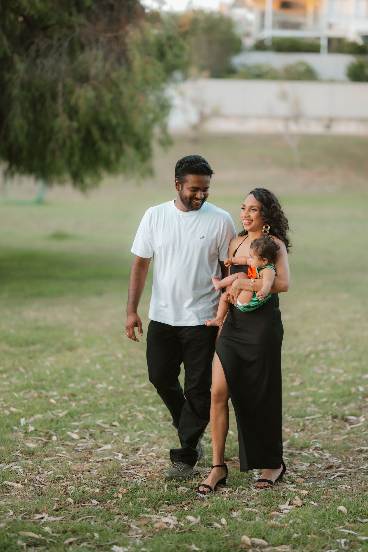 Happy couple in a park, man in white shirt and woman in black dress holding a baby, walking outdoors.