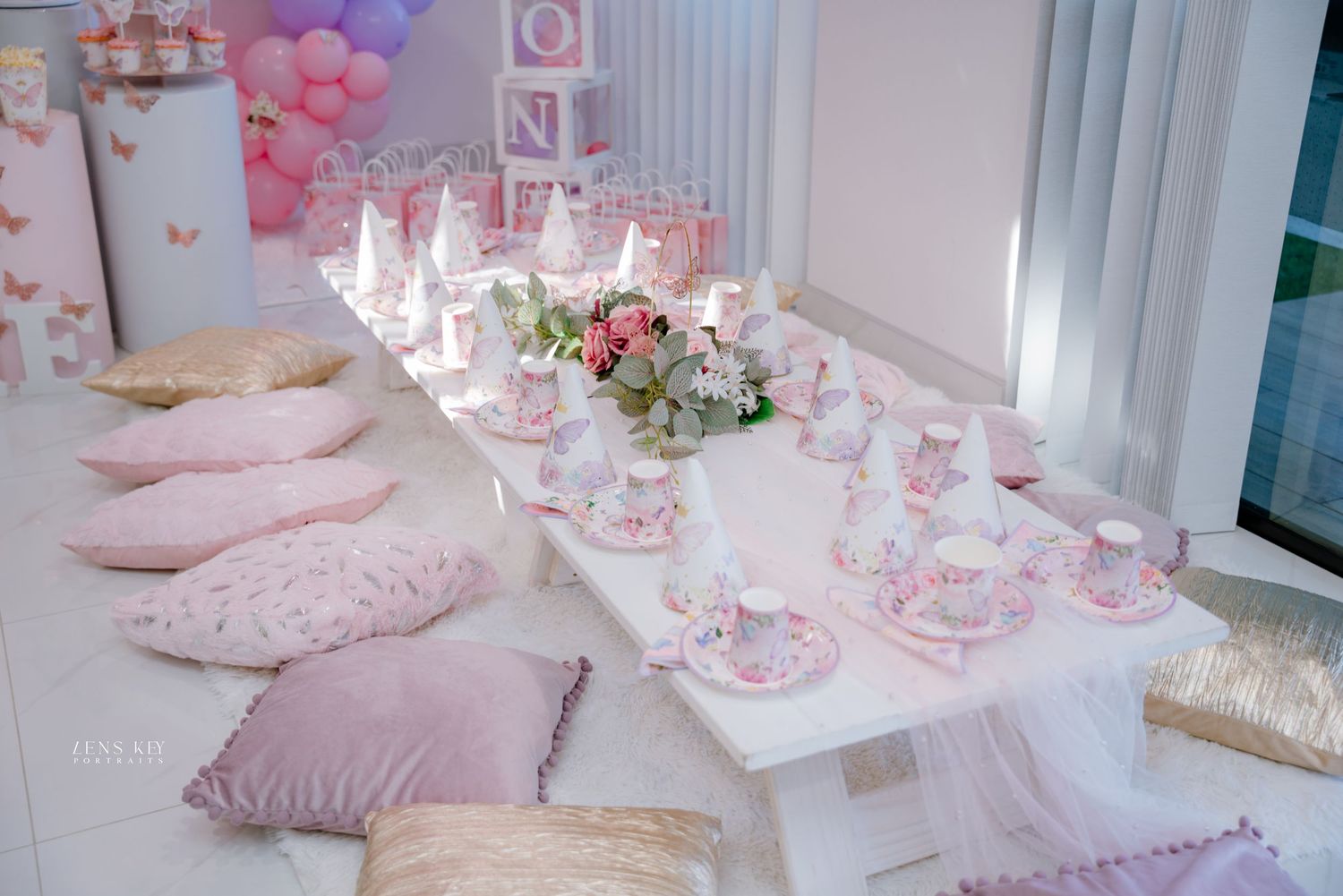Pink and purple butterfly birthday party table setting with low white table, cushions, and floral centerpiece.
