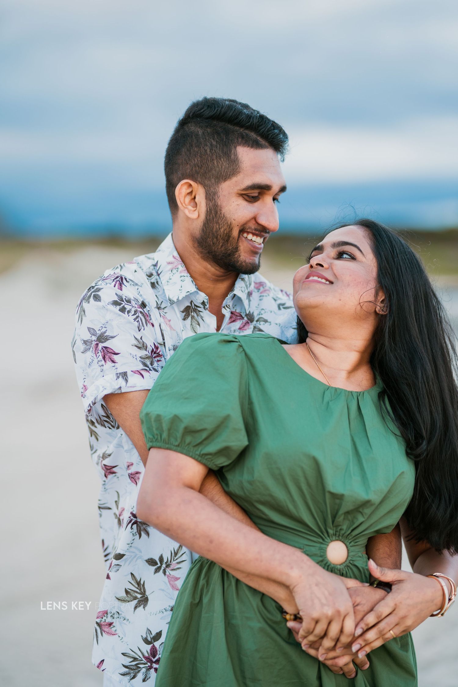 Couple embracing and smiling at each other on a beach, woman in green dress and man in floral shirt.