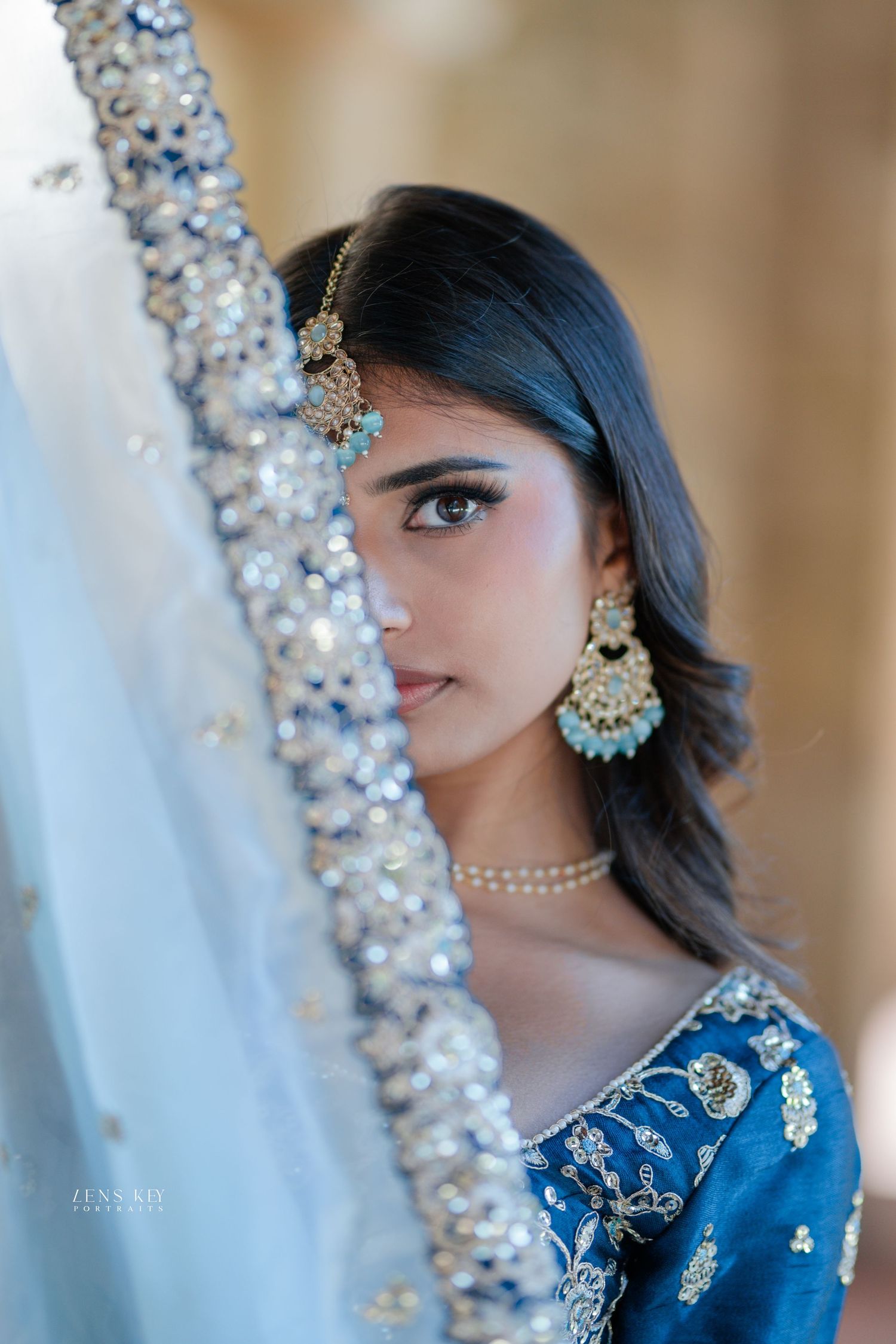 A bride in blue embroidered lehenga with kundan maang tikka and pastel chandbali earrings poses behind ornate dupatta.