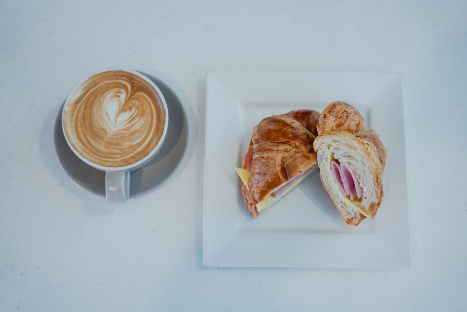 A latte with leaf art beside a ham and cheese croissant sandwich on a white plate.