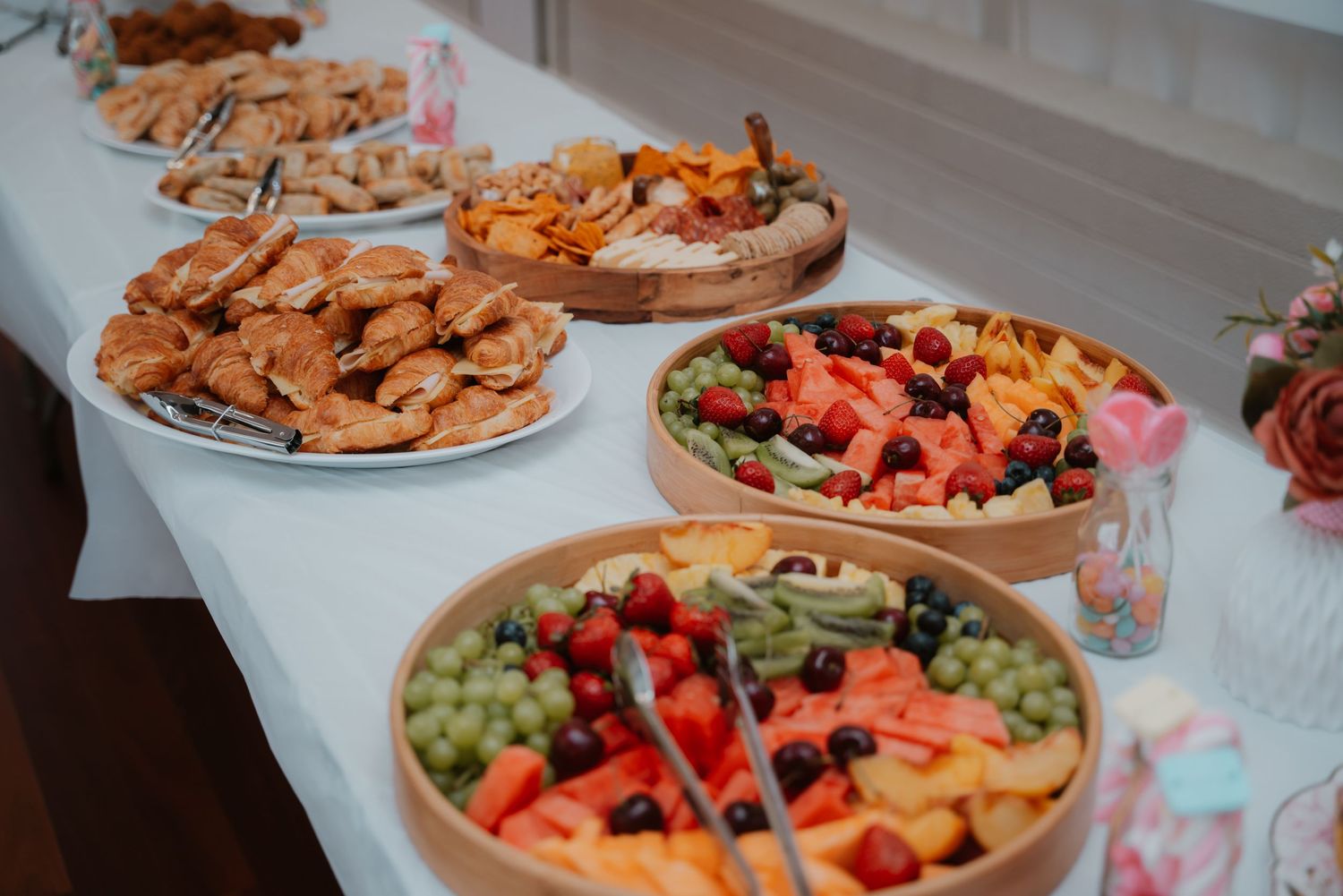 Colorful party food spread with fresh fruit platters and savory snacks on a white tablecloth.