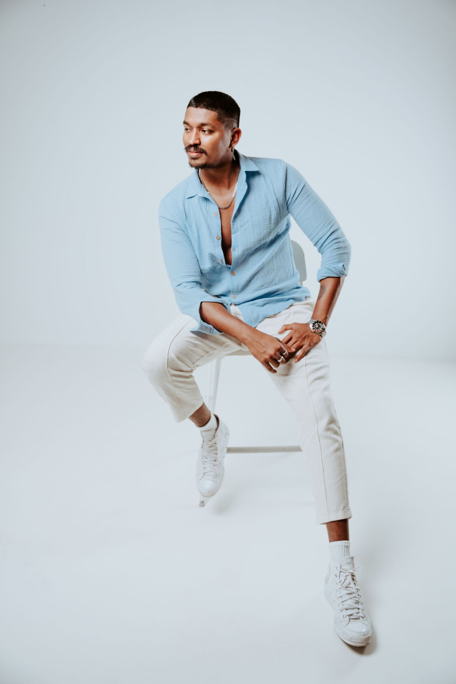 Confident man in light blue shirt and cream pants sits on stool against white background.