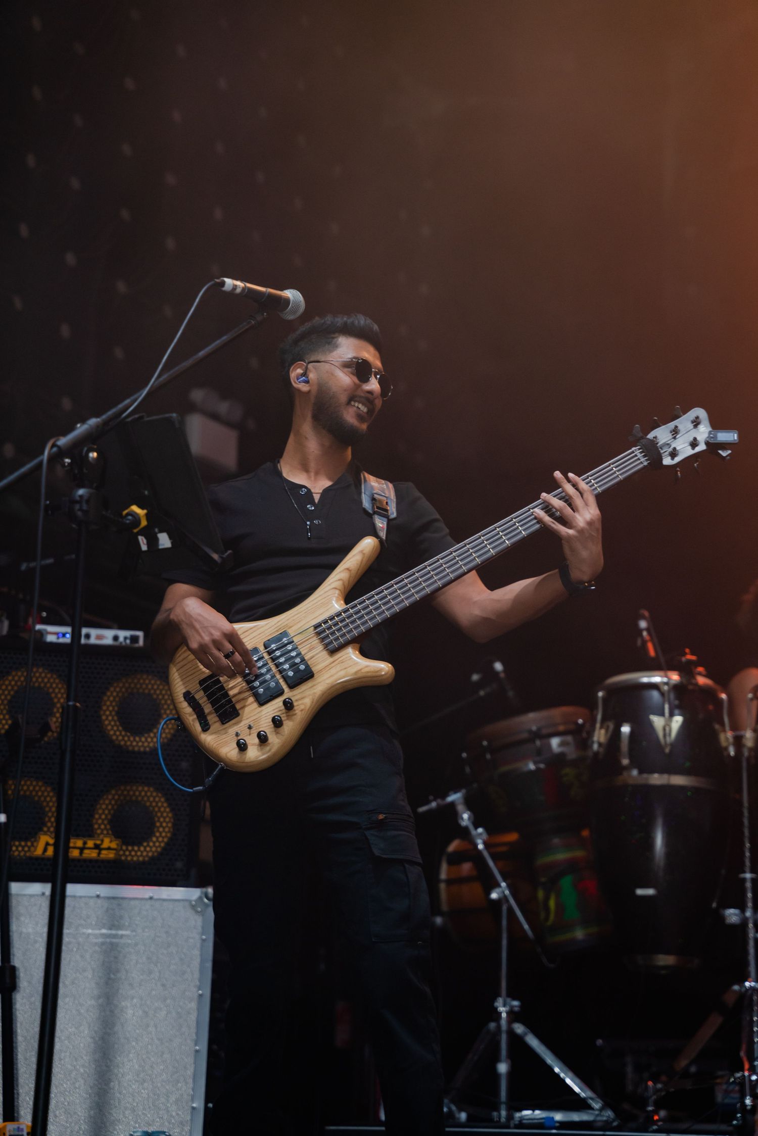 Male bassist in black outfit plays electric bass guitar on stage with warm amber lighting.