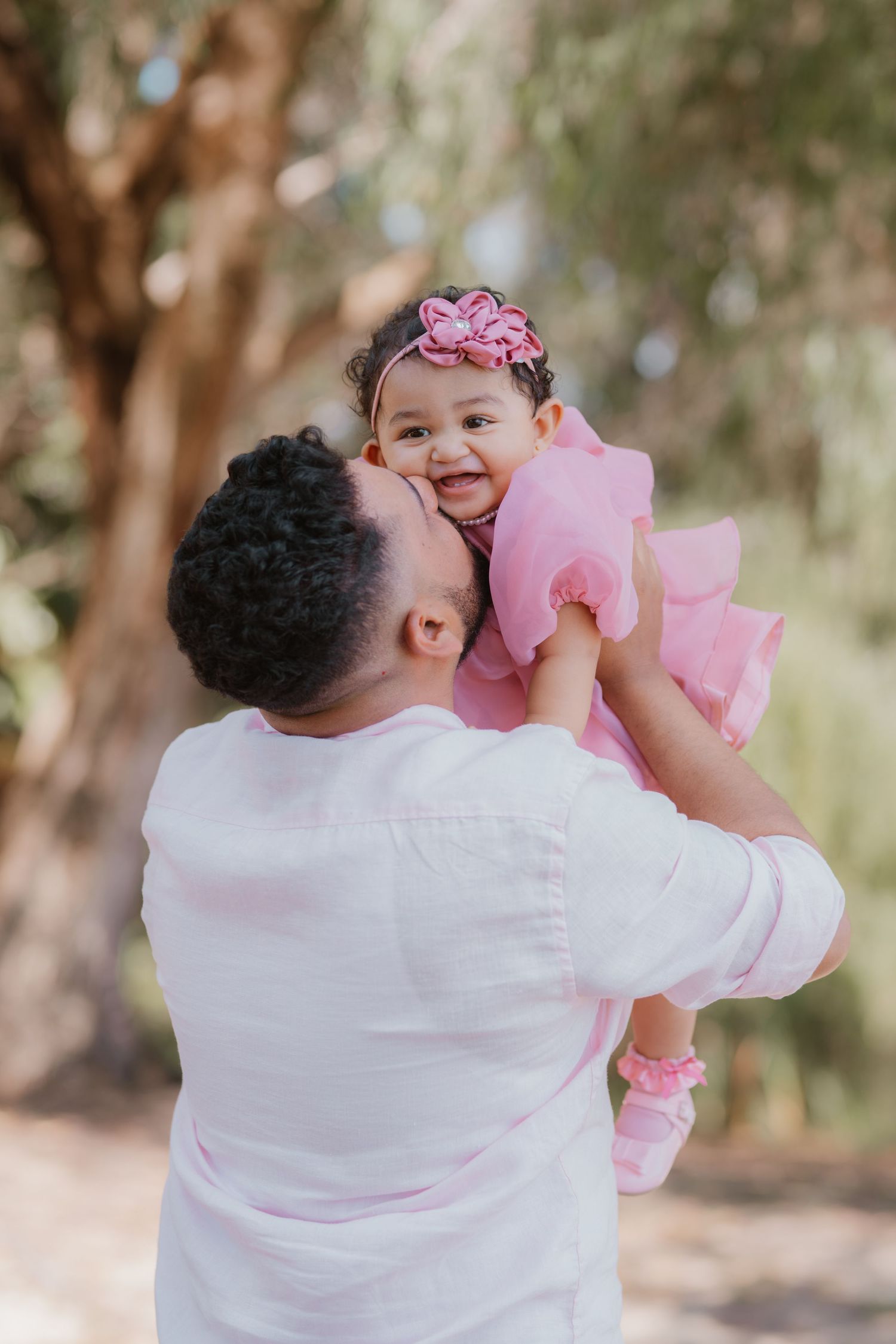 Father in white shirt lifts smiling baby girl in pink dress and floral headband outdoors.